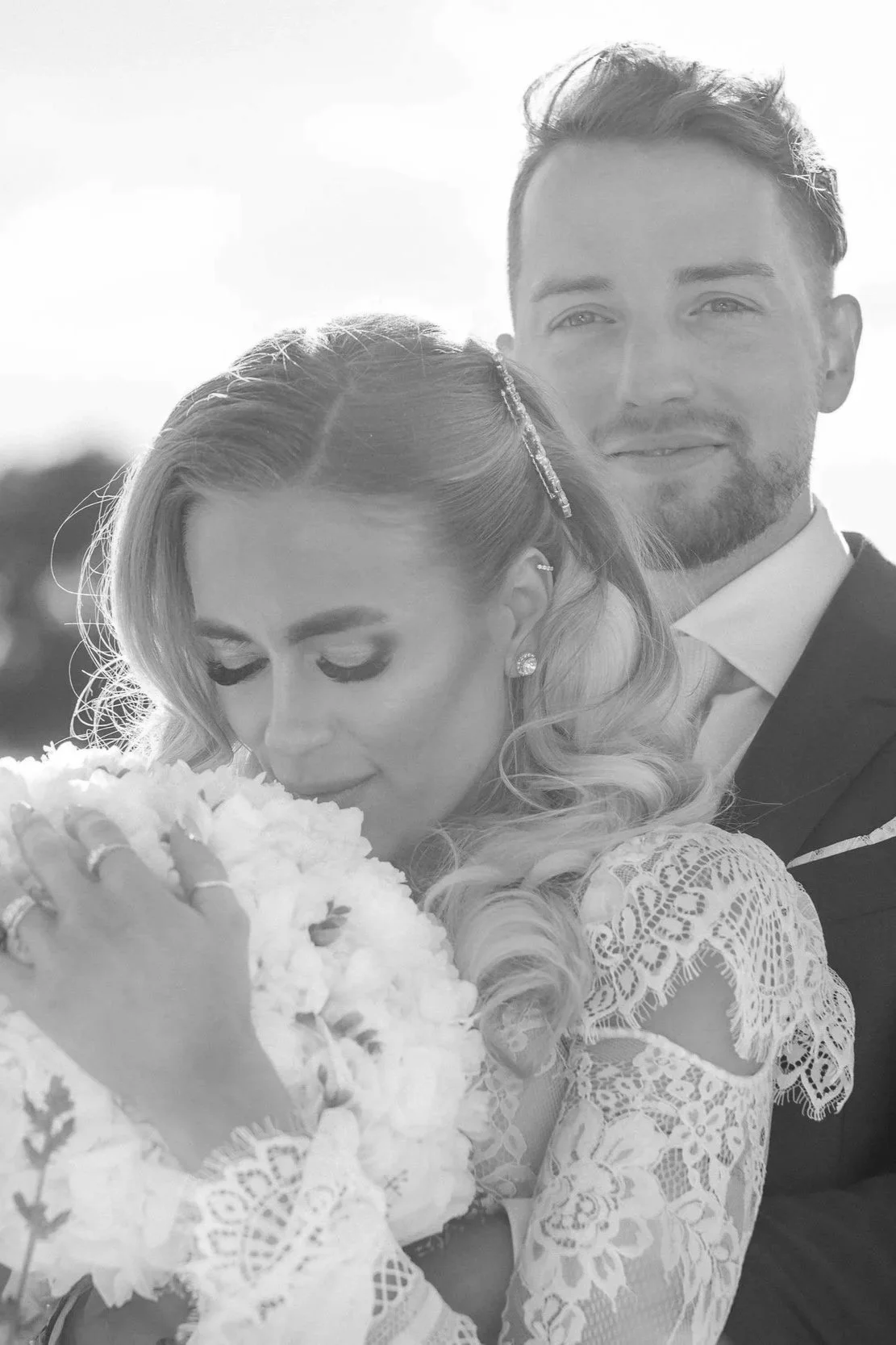 Black and white photo of a bride and groom on their wedding day, with the bride holding a large bouquet of flowers and the groom standing behind her, both smiling gently
