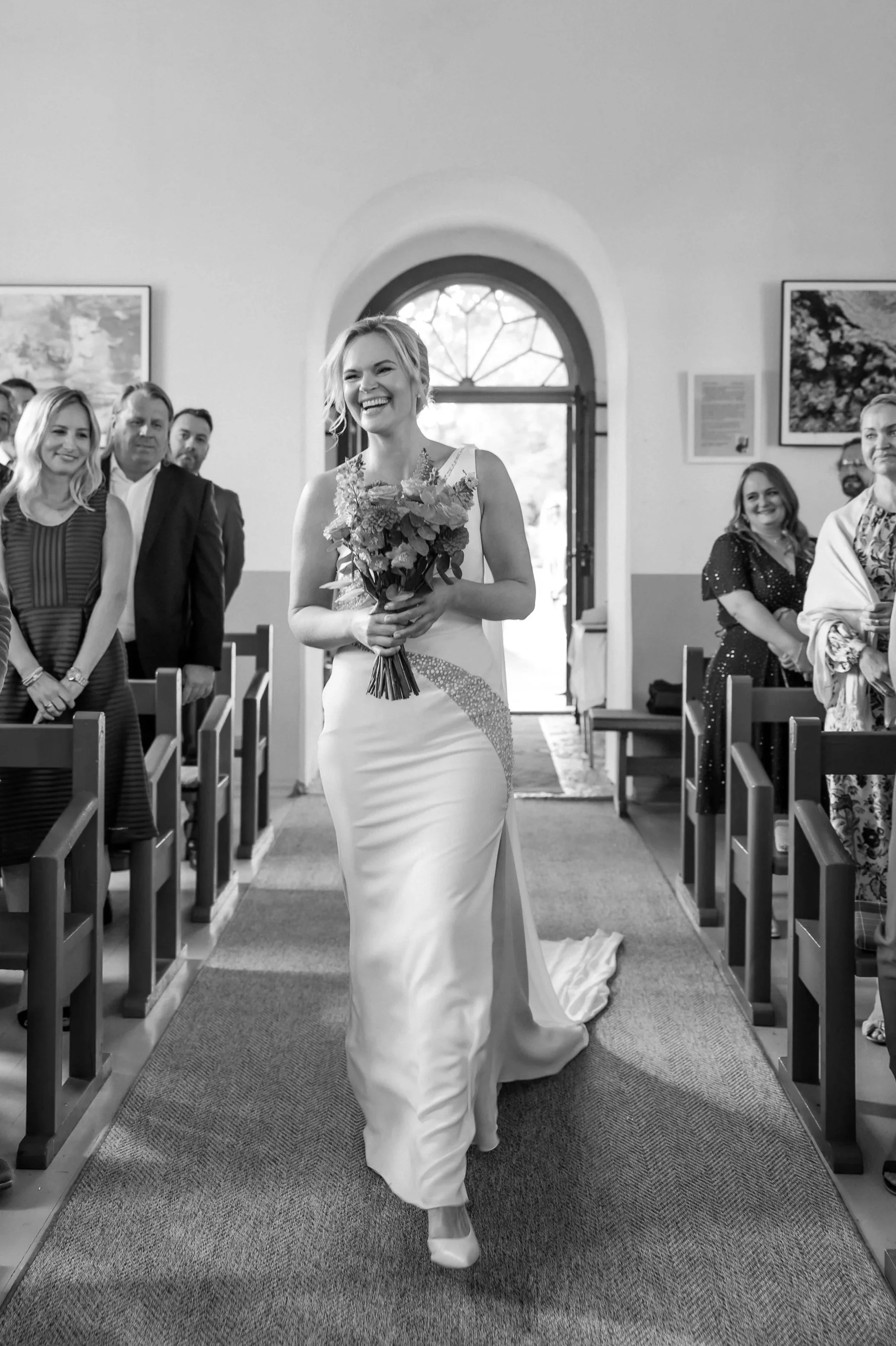 A woman in a wedding dress walking down the aisle of a church while holding a bouquet of flowers, with guests smiling and watching her.