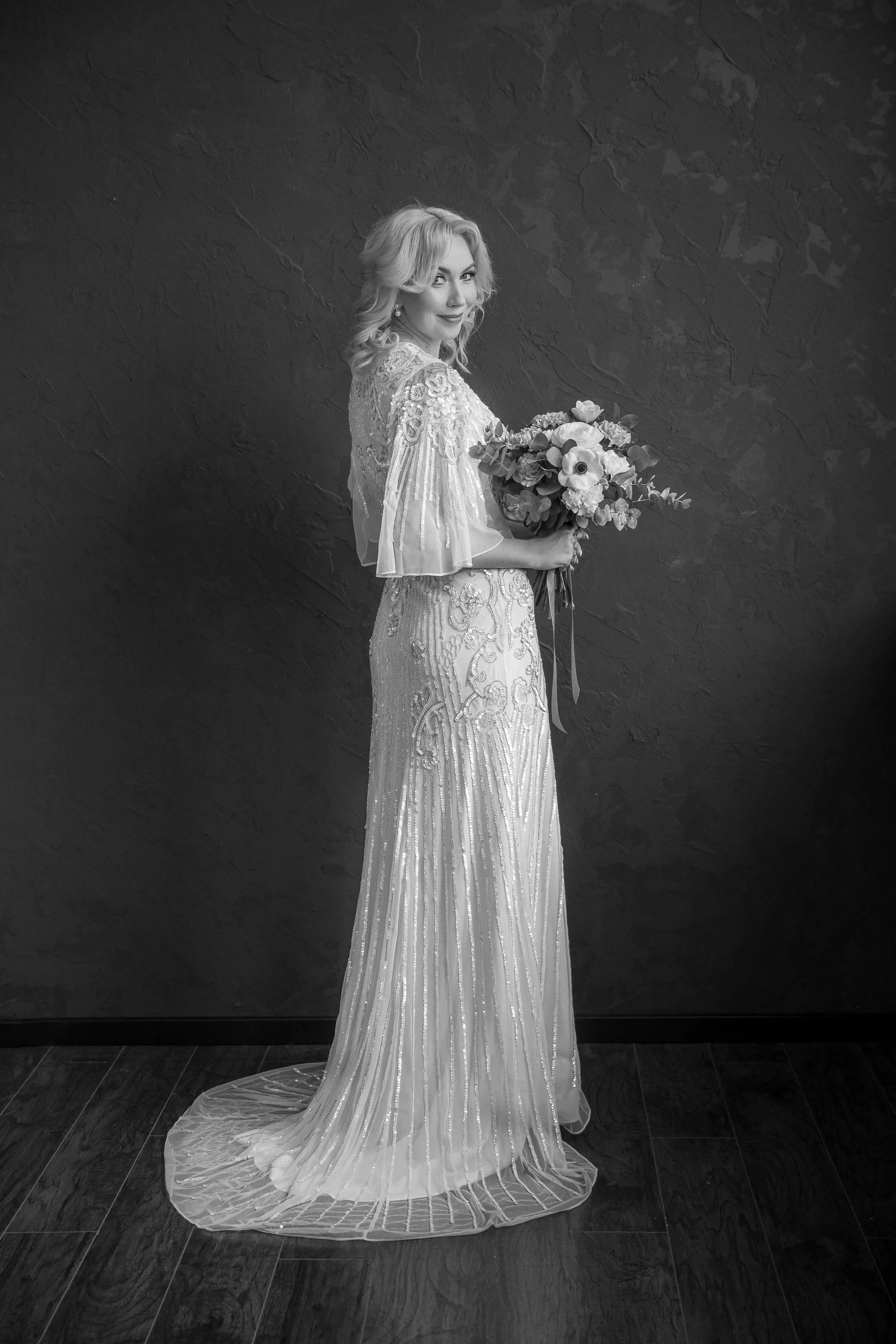A woman in a wedding dress holding a bouquet of flowers, standing against a dark textured wall.