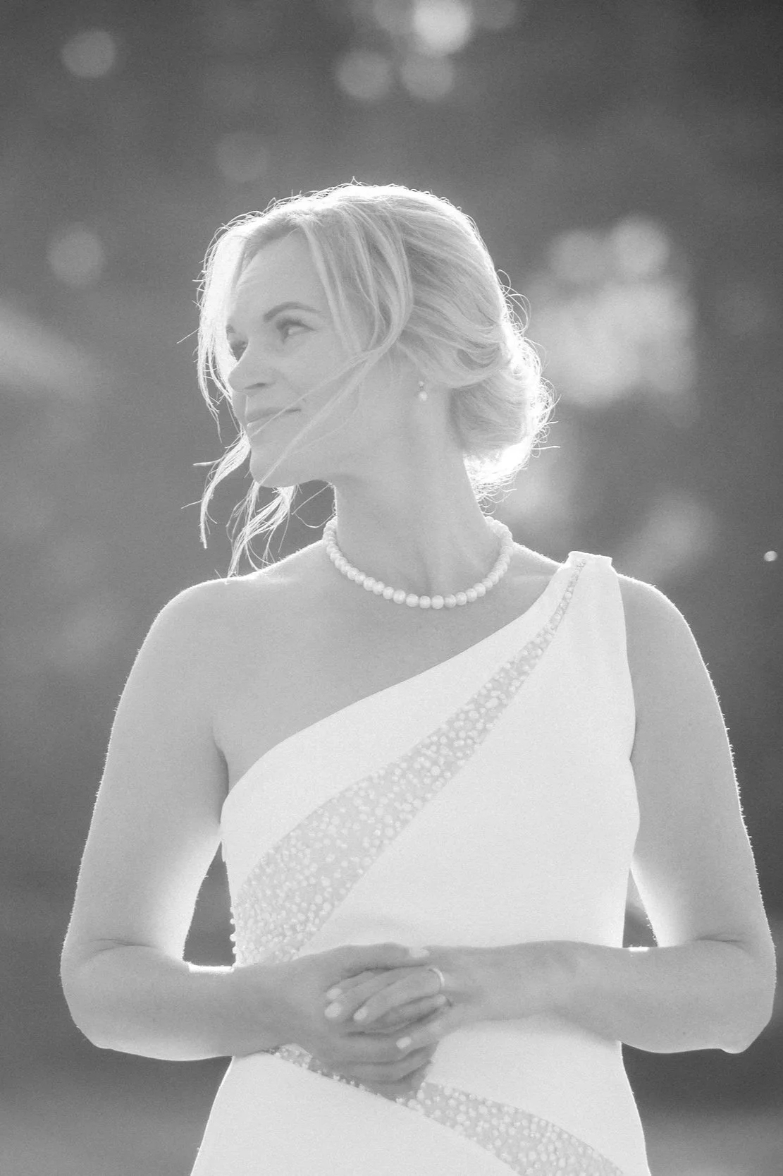 A woman in a white dress with a pearl necklace stands outdoors with blurred lights in the background, looking to the side.