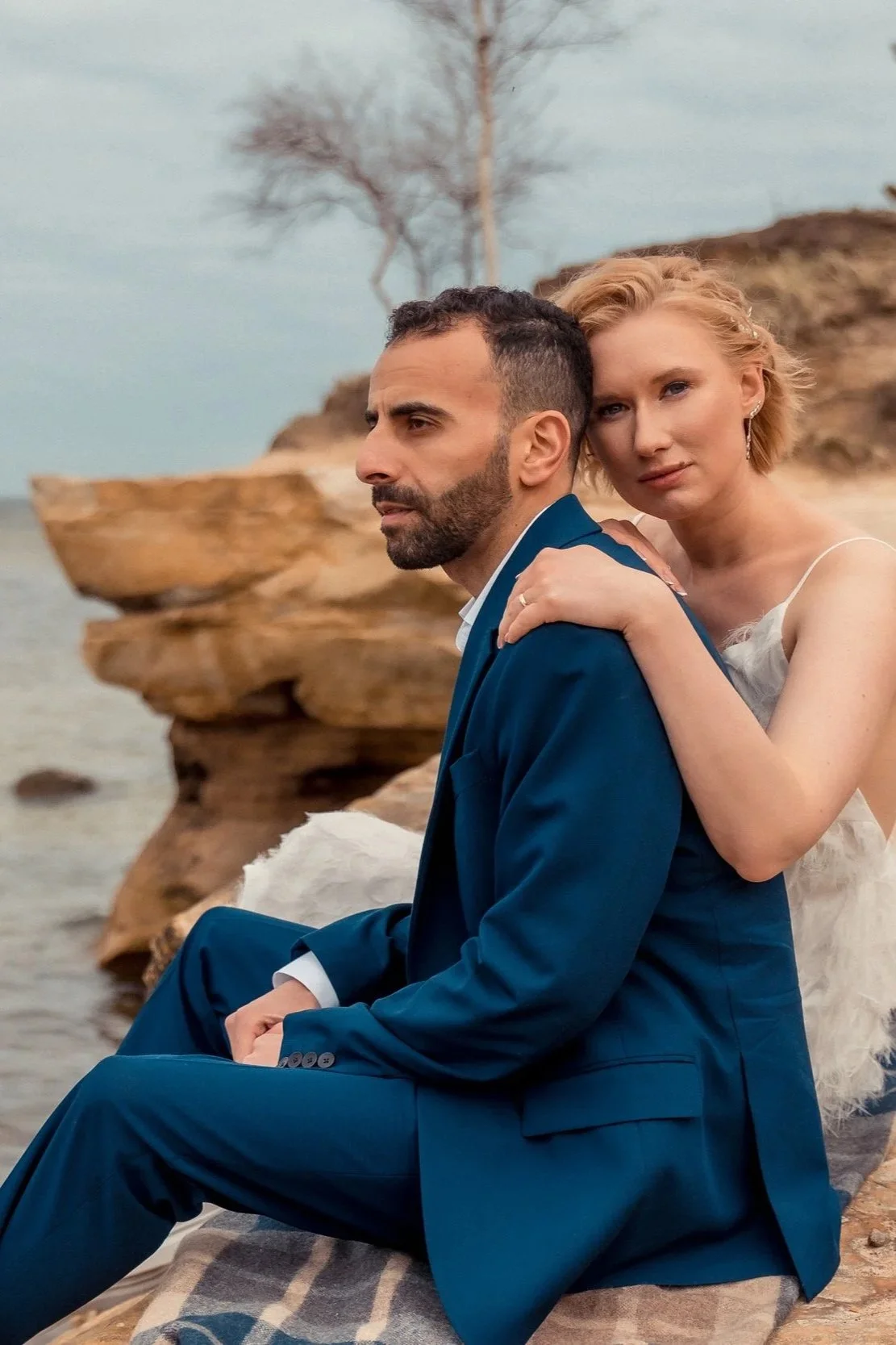 A man in a blue suit and a woman in a white dress sitting by the water near rocks, with the woman embracing the man from behind in an outdoor setting.