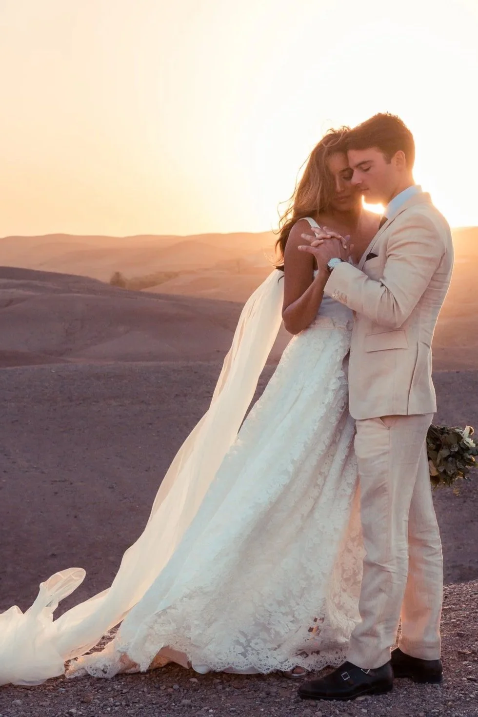 A couple dressed in wedding attire, embracing outdoors at sunset with a barren landscape background.