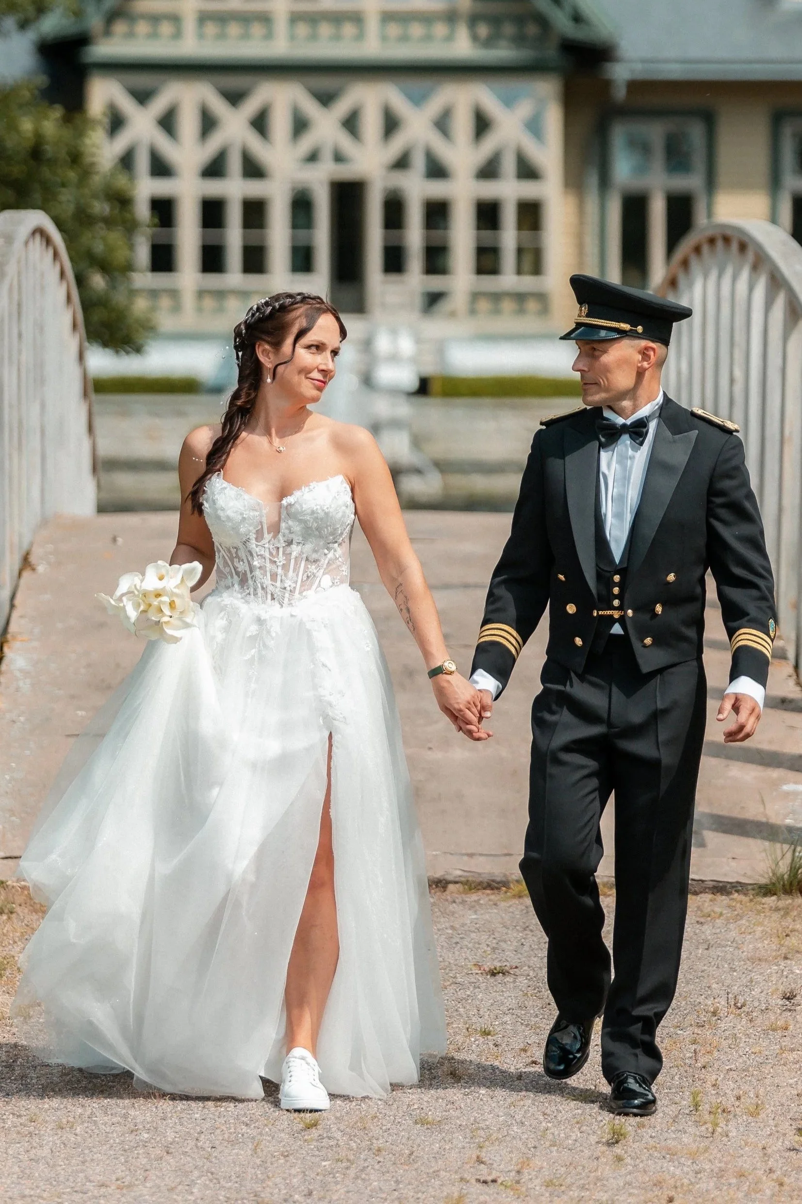 A bride and groom walking hand in hand outdoors near a historic building, with the bride in a white wedding dress and the groom in a military uniform.