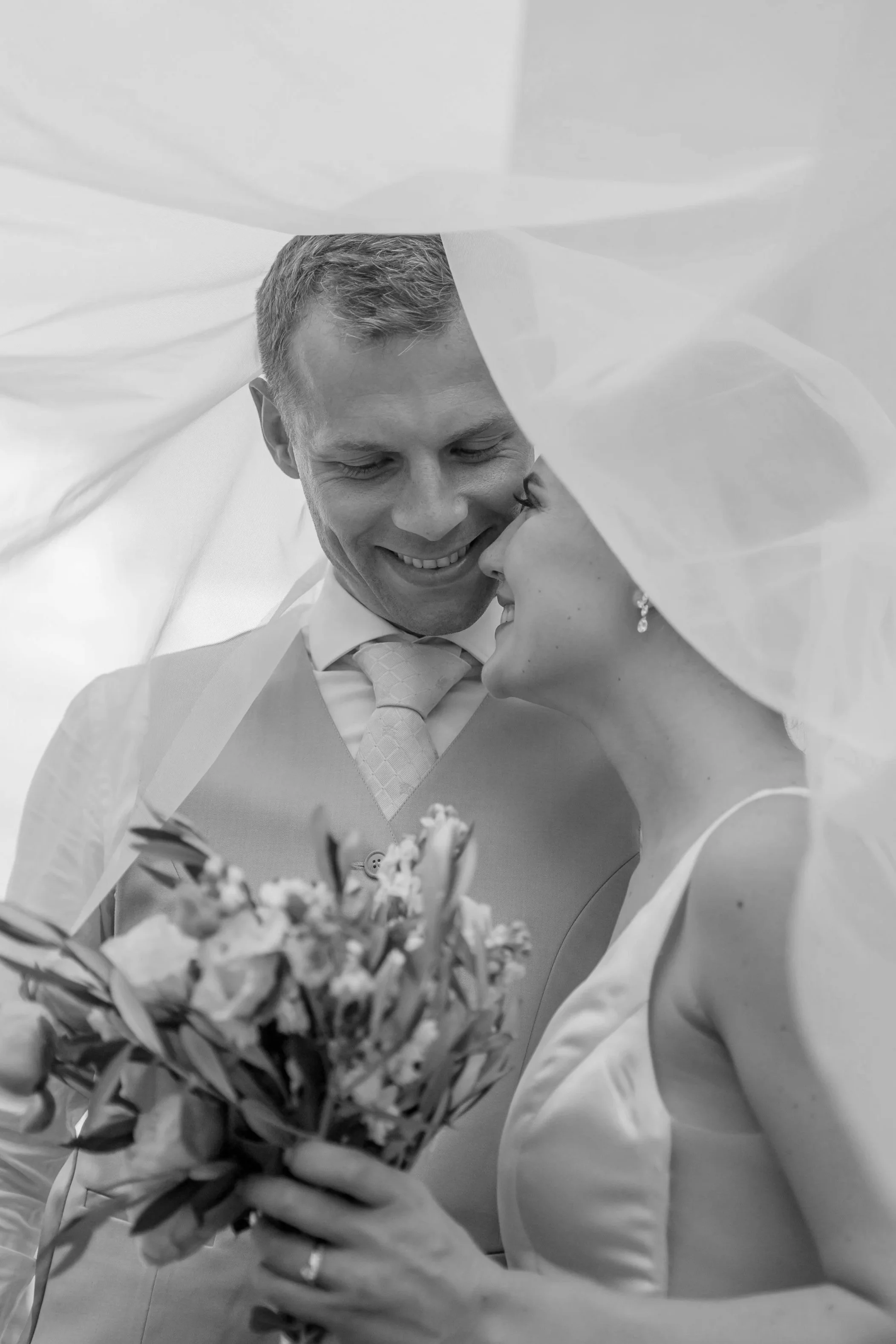 Black and white photo of a bride and groom smiling in an embrace, holding a bouquet of flowers.