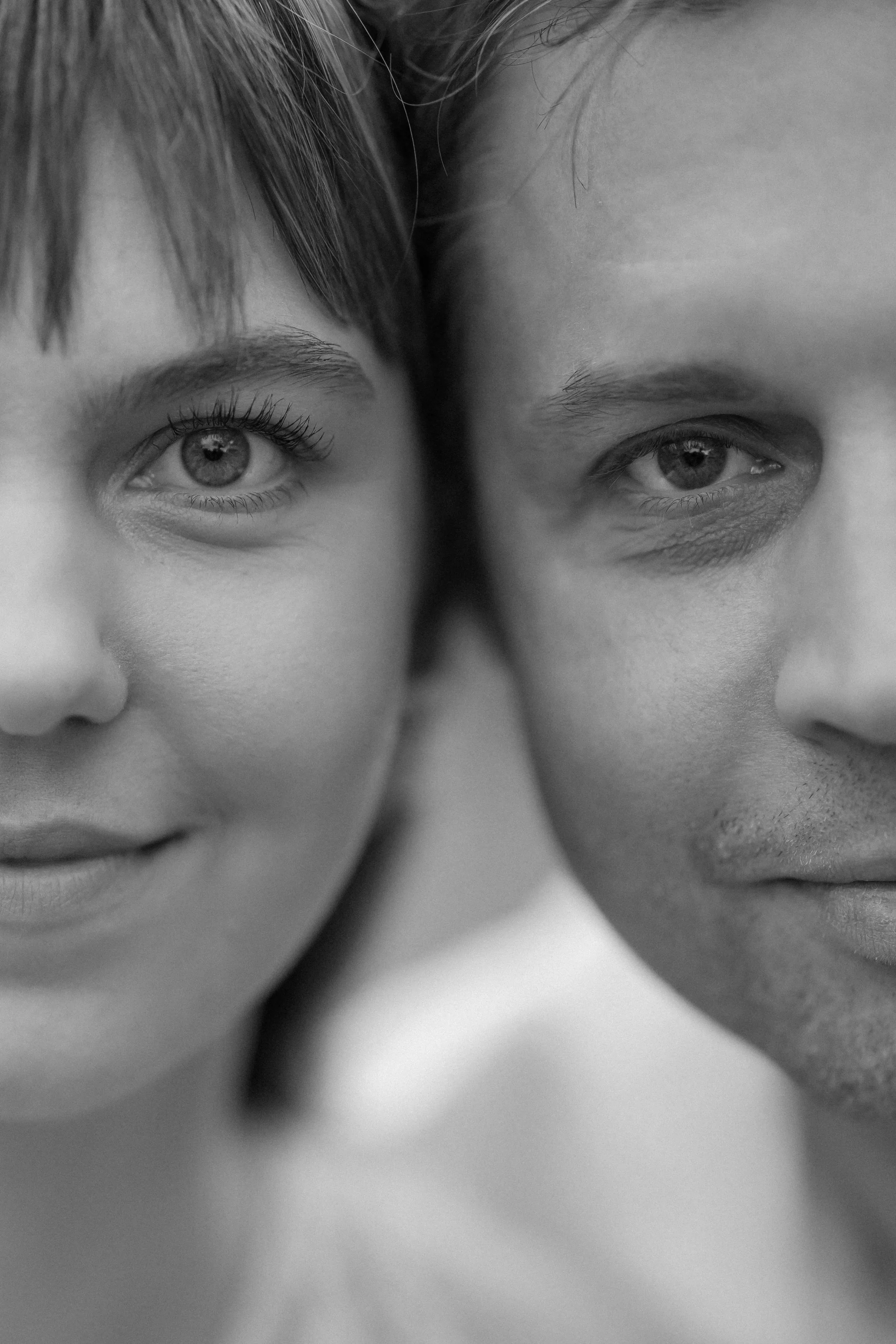 Black and white close-up photo of two faces touching cheeks, with one face on the left side and the other on the right side, both smiling subtly.