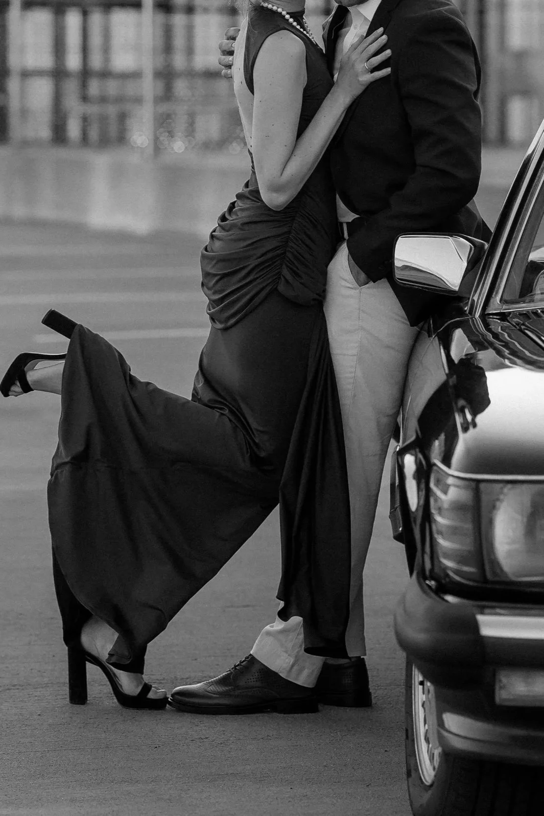 A black and white photo showing a woman kneeling on one knee with one leg extended, wearing high heels, and embracing a man who is leaning against a car. The woman is dressed in a gown and jewelry, and the man is wearing a blazer, dress shirt, and pa