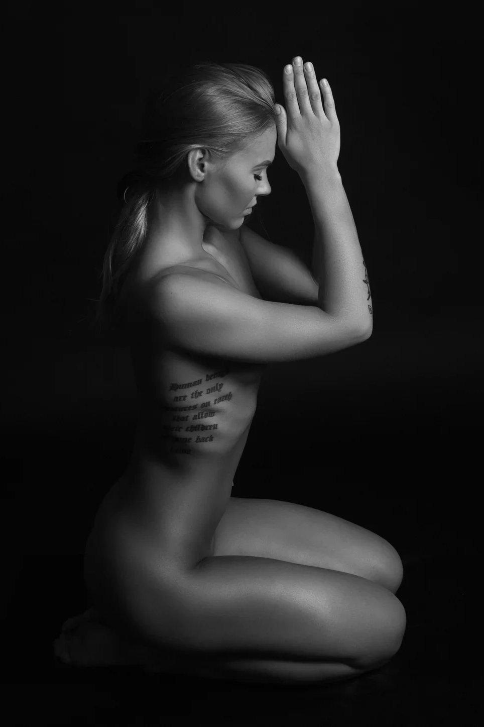 A woman kneels with her hands pressed together in a prayer position, eyes closed, against a black background. She is nude, with long hair, and has tattoos on her side and arm.
