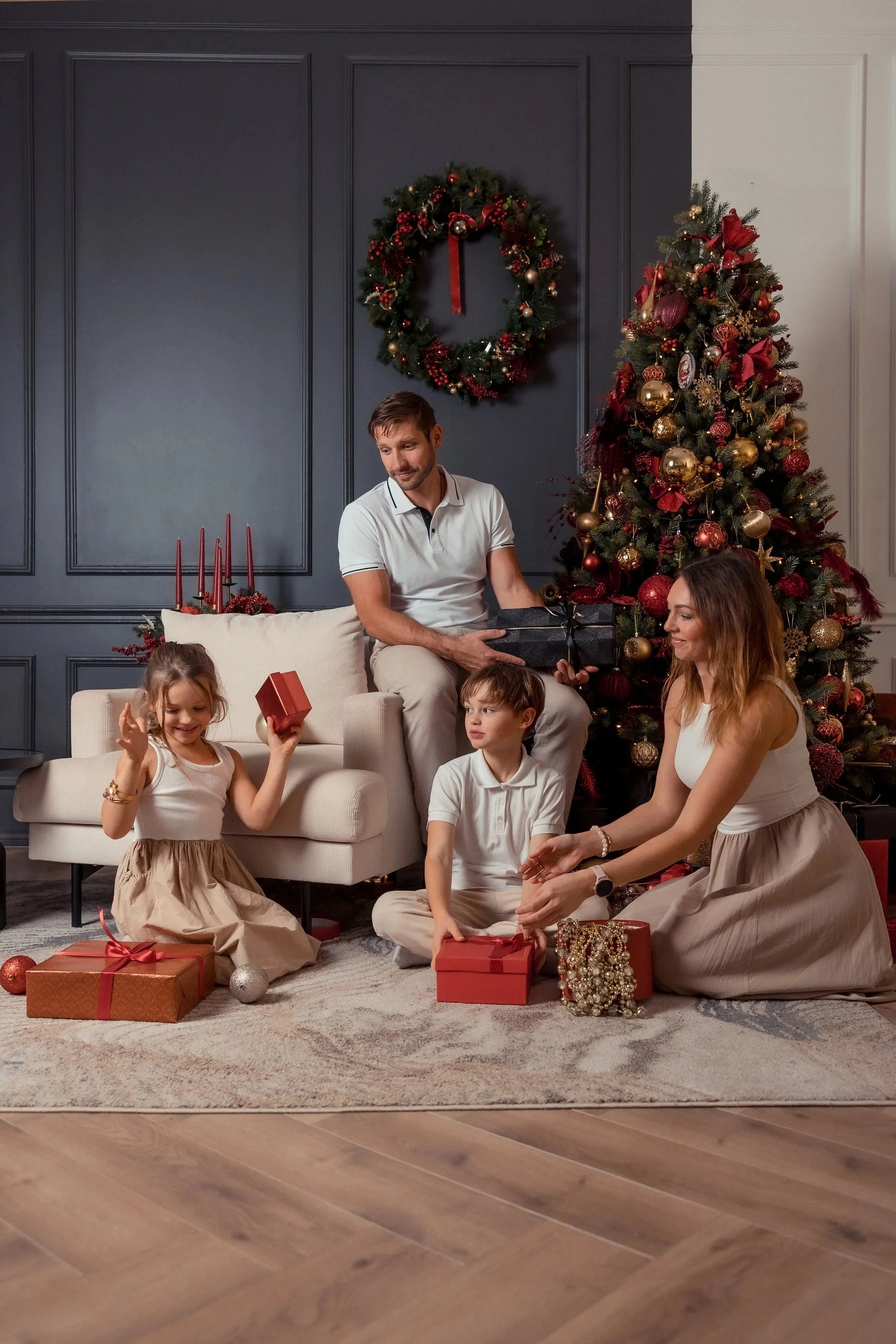 A family celebrating Christmas with a decorated tree, presents, and a wreath on a dark blue wall. The scene includes two children, a woman, and a man, all engaging in gift exchange and smiling.