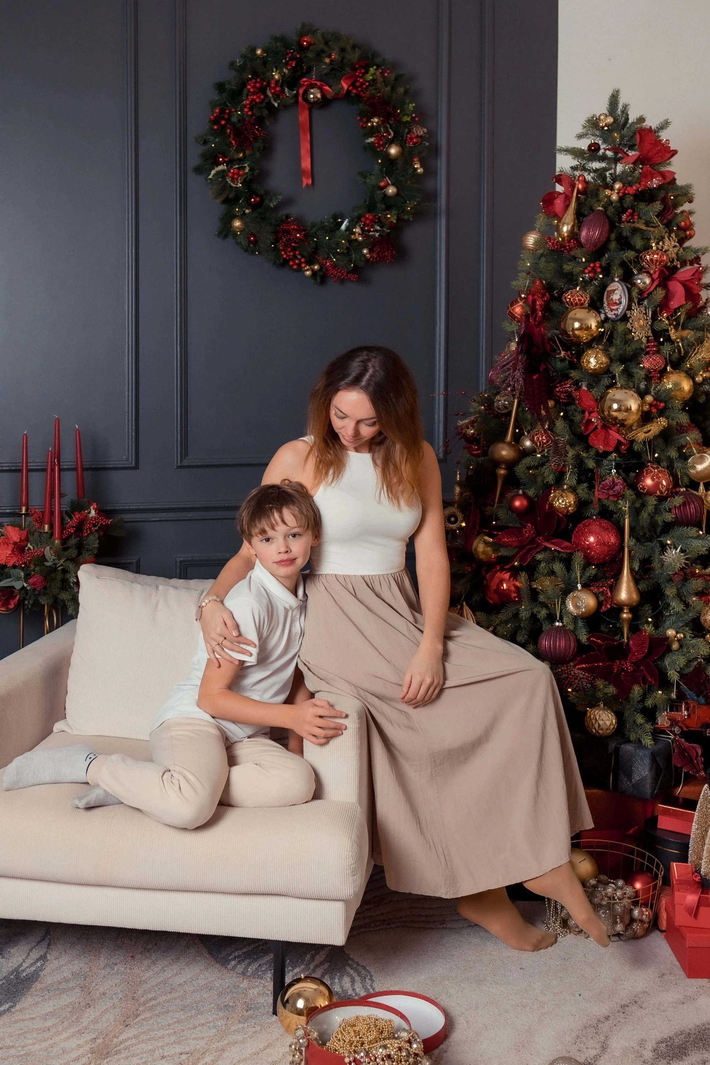 A woman and a young boy sitting on a cream-colored sofa in a festive living room. The woman is wearing a sleeveless white top and a beige skirt, sitting with her legs crossed. The boy is sitting on the sofa with his legs bent, wearing a white shirt a