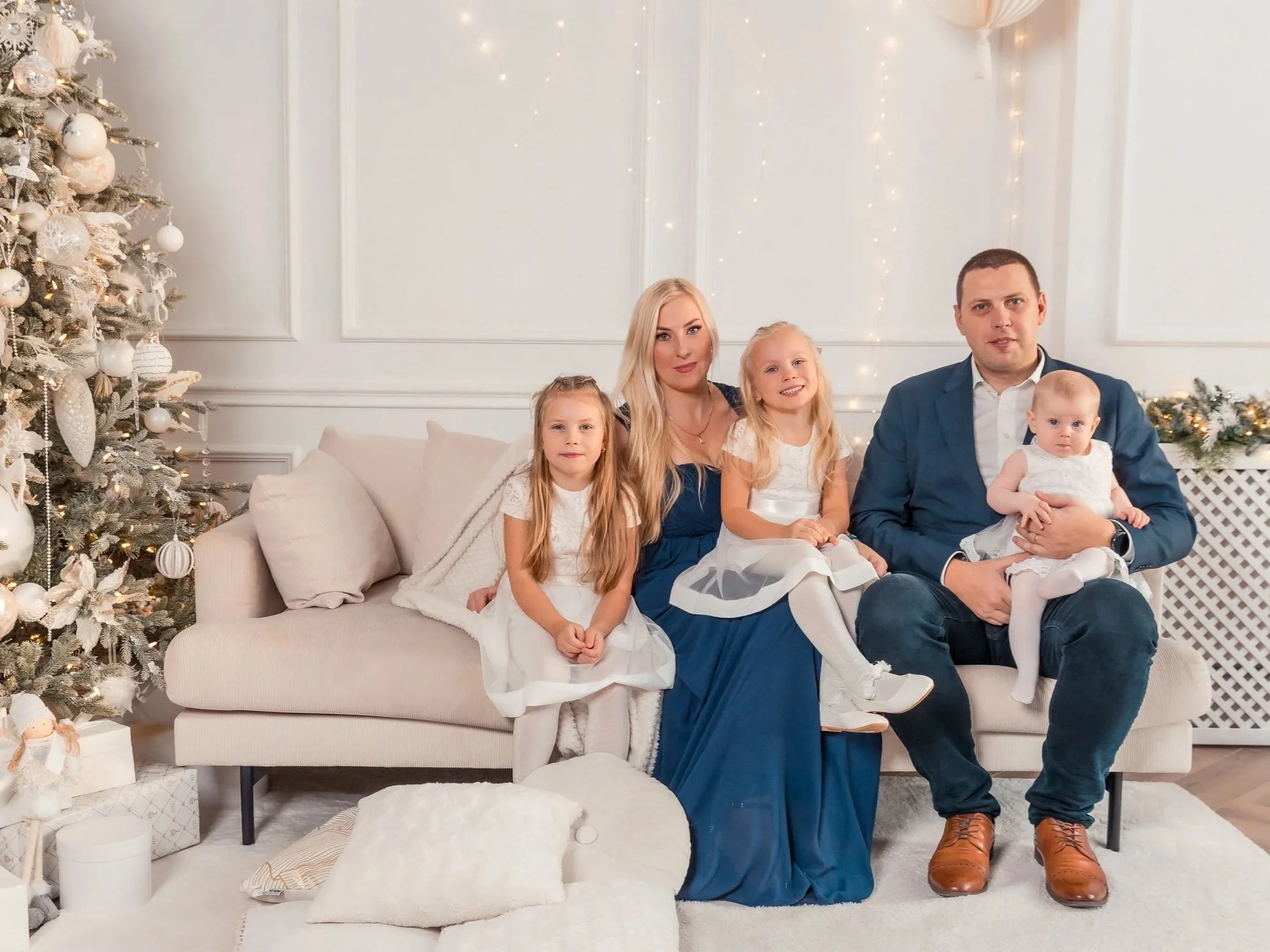 A family of five sitting on a beige sofa in a Christmas decorated room, with a Christmas tree on the left and fairy lights on the wall in the background.