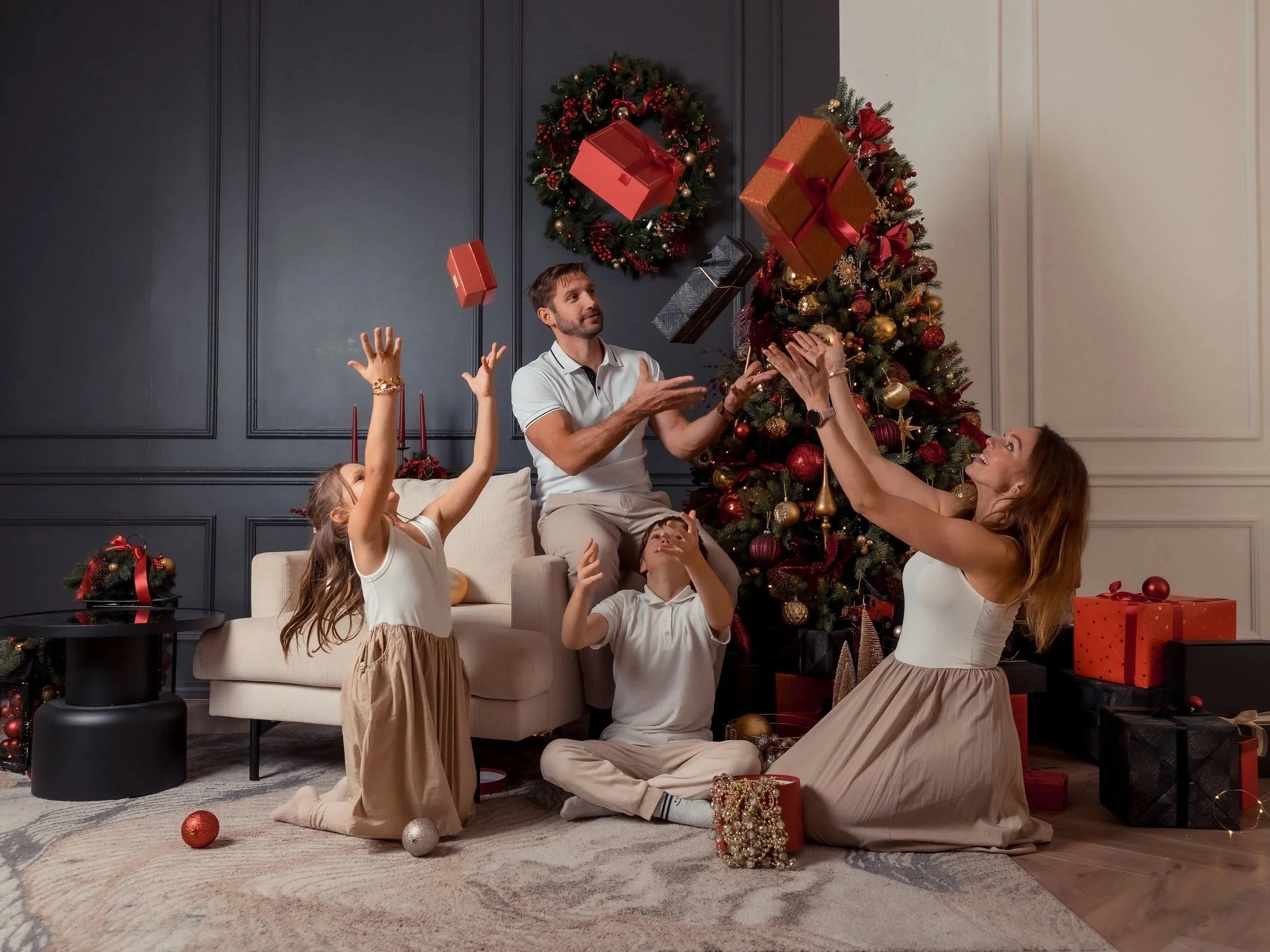 Family celebrating Christmas in living room with decorated Christmas tree, presents, and holiday decor, some family members are tossing gift boxes in the air.