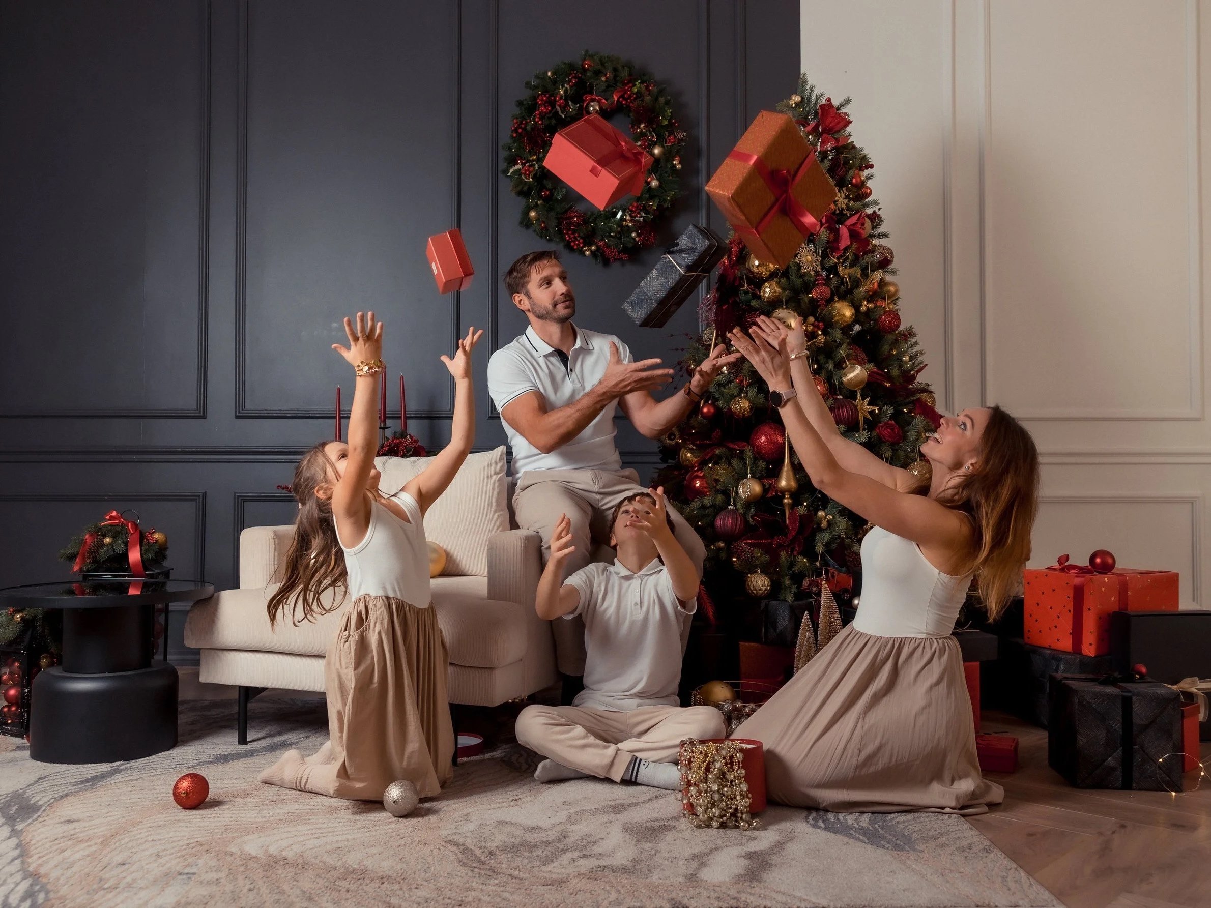 Family celebrating Christmas in living room with decorated Christmas tree, presents, and holiday decor, some family members are tossing gift boxes in the air.