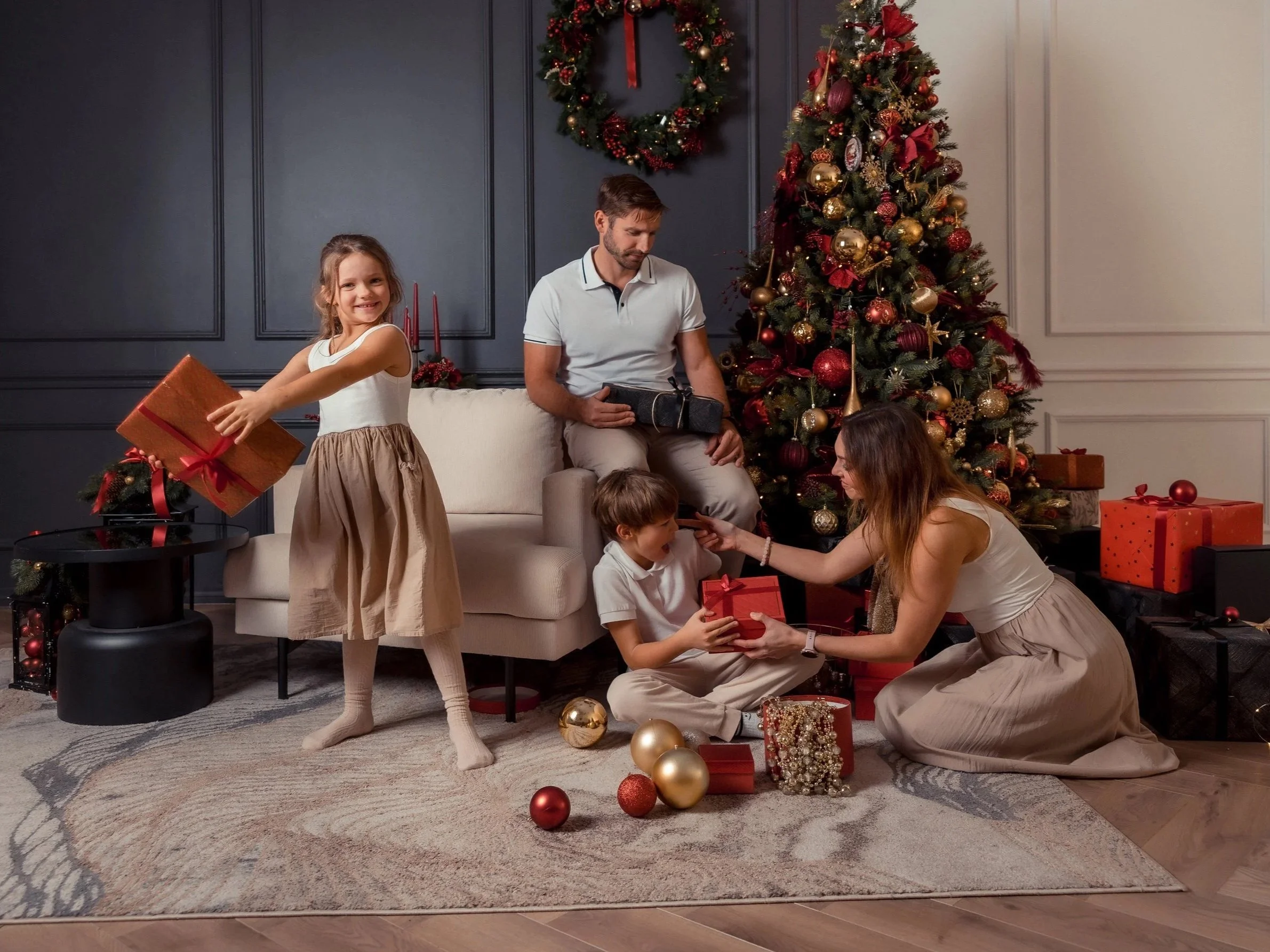 A family celebrating Christmas in a decorated living room. A Christmas tree with gold and red ornaments, a wreath hanging on the wall, and wrapped presents are visible. A young girl with brown hair is holding a gift, while a woman and two children ar