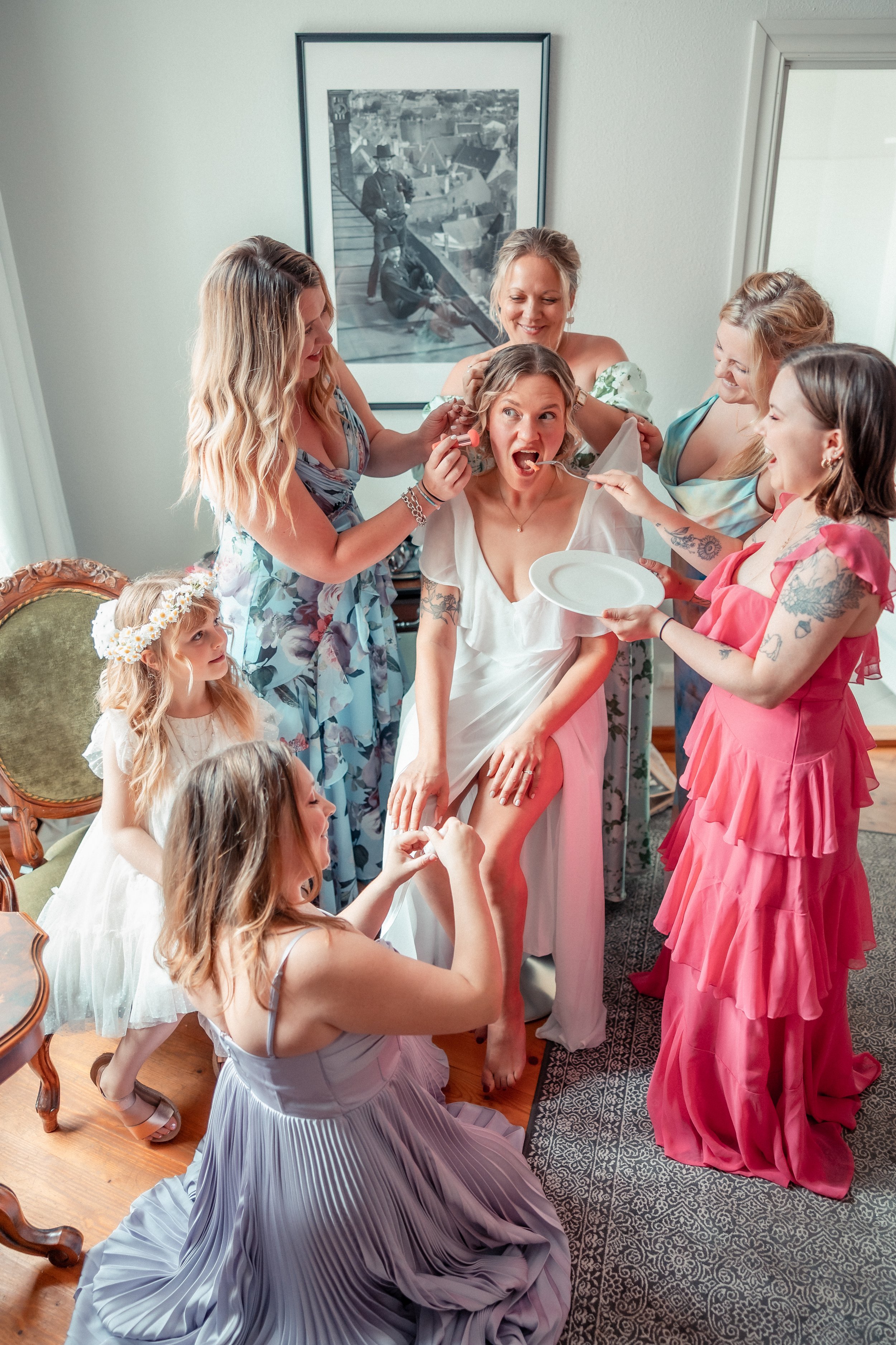 A bride in a white dress having makeup or face paint applied by women and girls surrounding her in a room with floral decor and art on the walls.