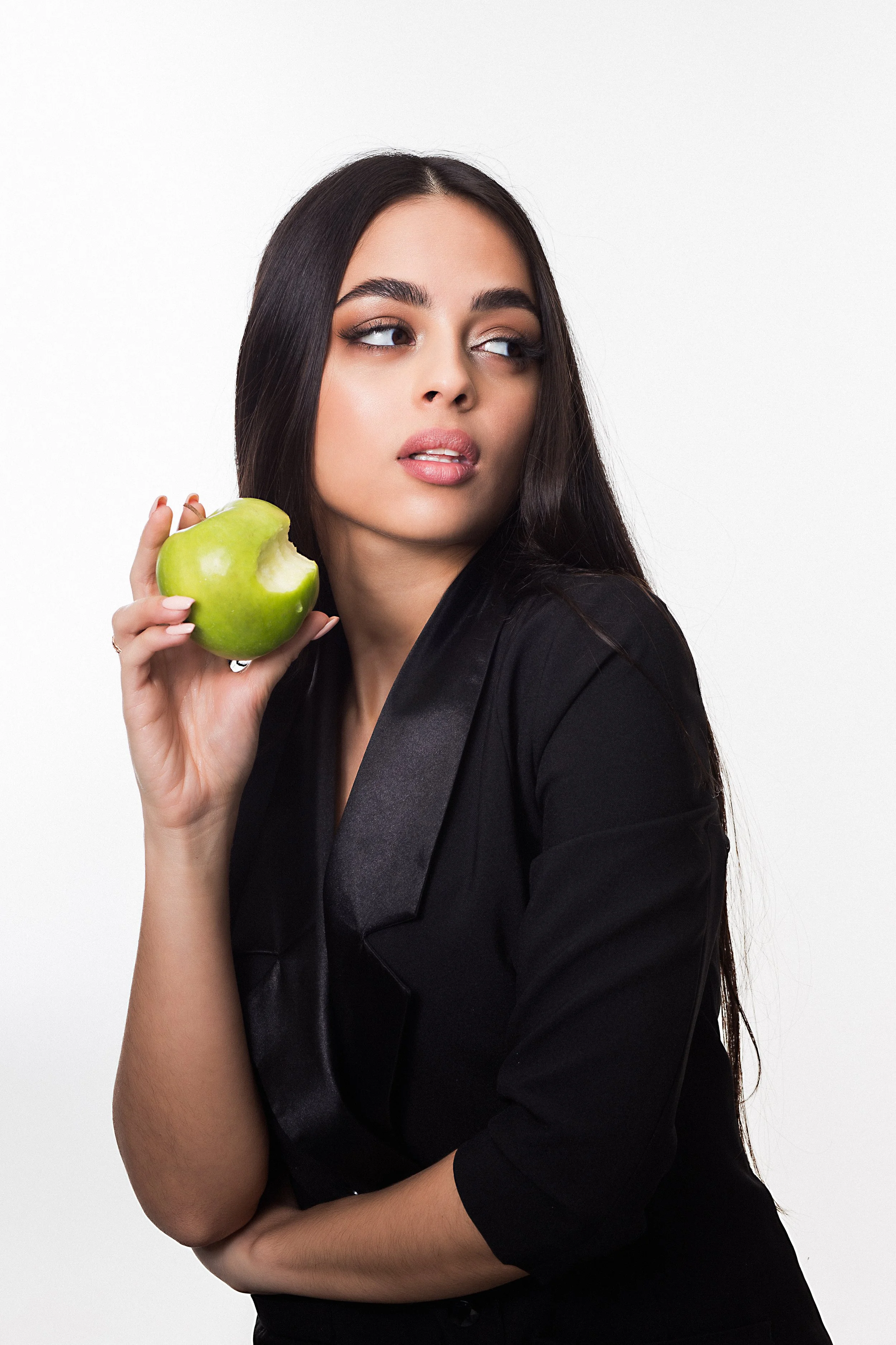 A young woman with long dark hair and makeup, holding a partially eaten green apple, wearing a black outfit, against a plain white background.