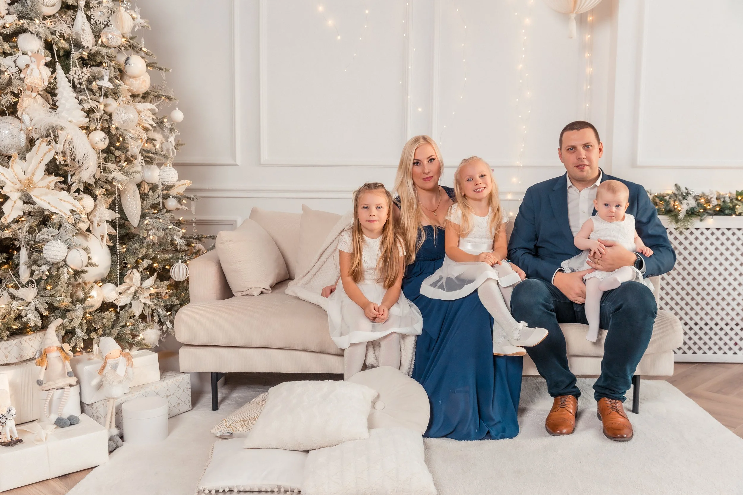 A family of five sitting on a cream-colored sofa next to a decorated Christmas tree in a festive living room. The two young girls are dressed in white and blue dresses, the woman has long blonde hair, and the man is wearing a blue suit jacket. The yo