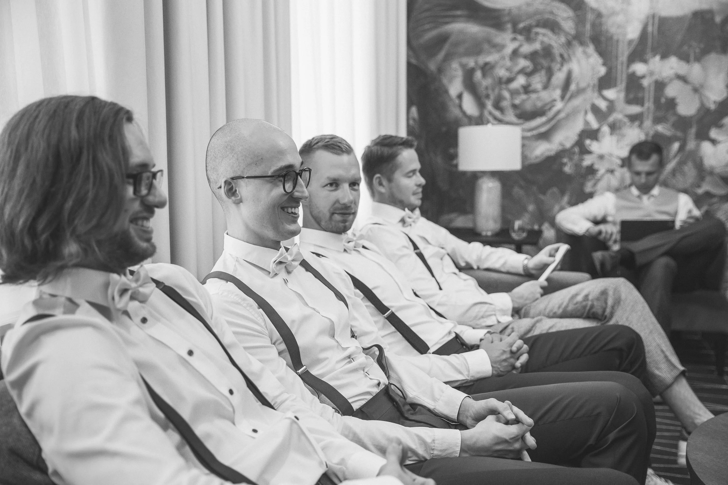 A group of five men dressed in white shirts, bow ties, and suspenders, sitting in a row at a formal event, smiling and engaging with their phones, with a large floral painting and curtains in the background.