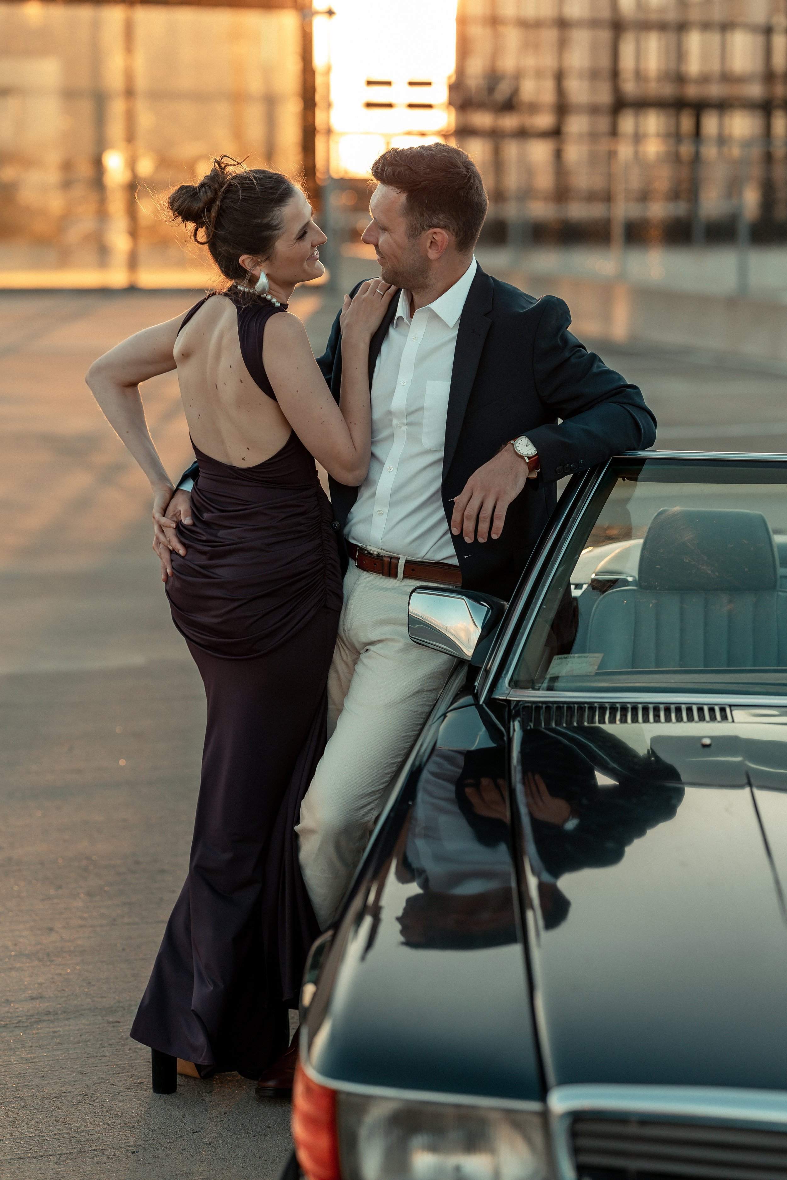 A woman in a black dress and pearl necklace leaning towards a man in a white shirt and blazer, standing next to a parked car on a rooftop parking lot during sunset.