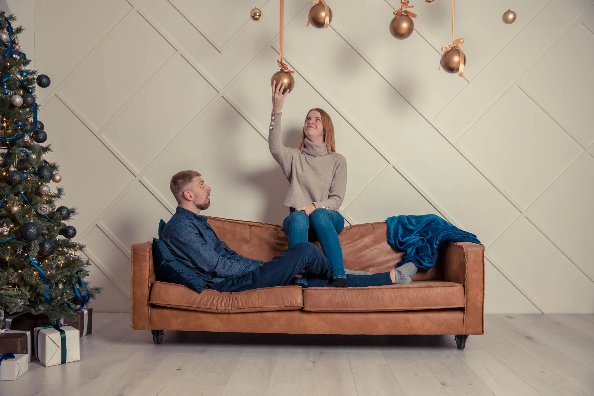 A woman sitting on a brown sofa, reaching up to touch hanging gold Christmas ornaments, while a man sits on the sofa looking up at her. There is a decorated Christmas tree with blue ribbons and ornaments to the left, and wrapped presents underneath. 