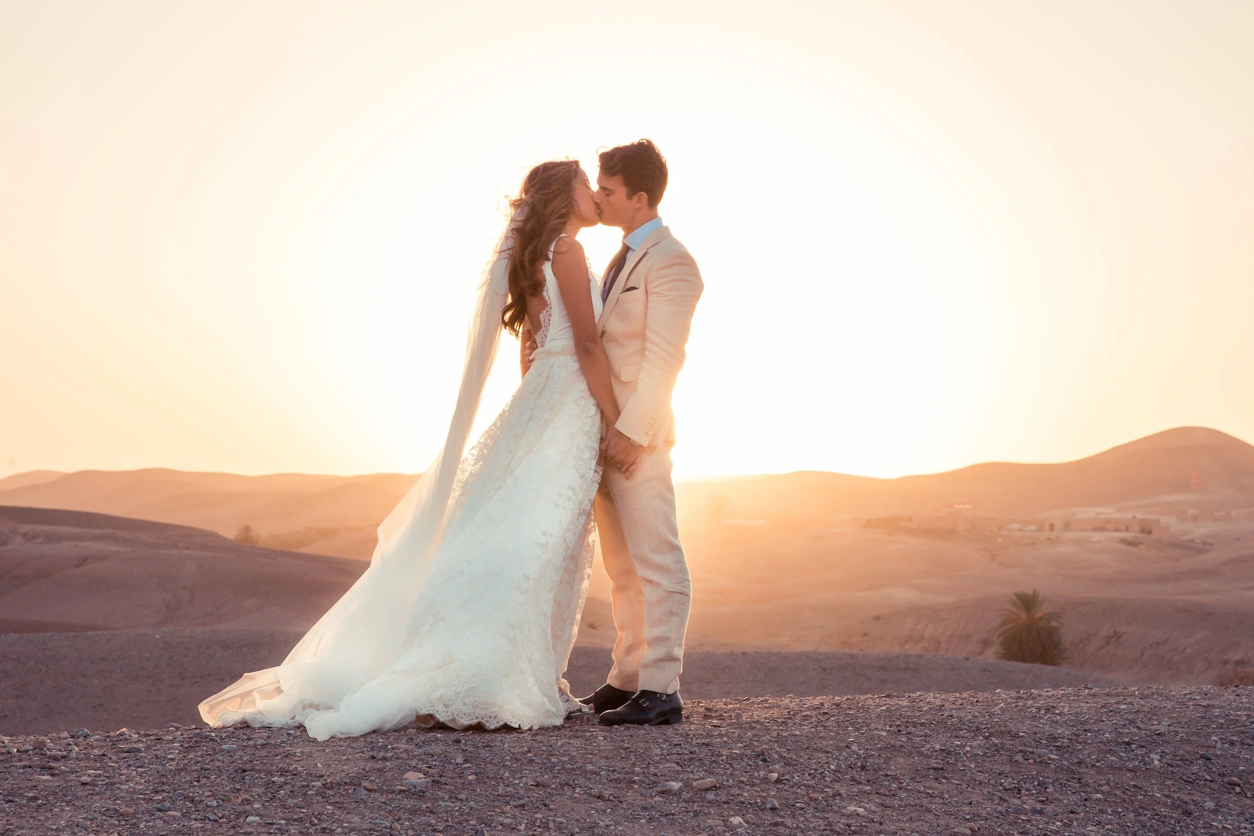 A couple in wedding attire kissing during sunset in a desert landscape.