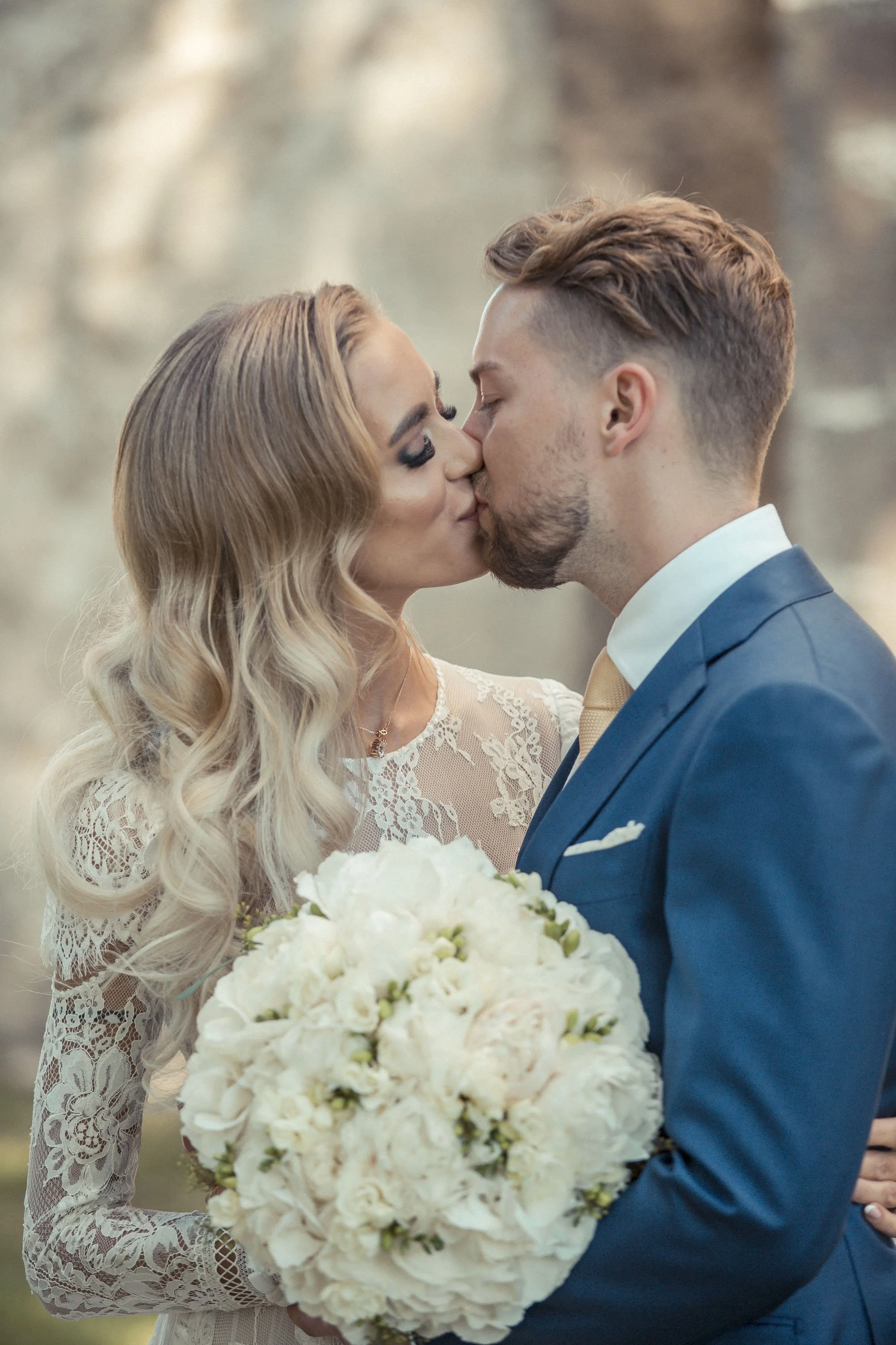 A bride and groom sharing a kiss during their wedding, with the bride holding a large bouquet of white flowers, and the bride wearing a lace wedding dress with long sleeves.