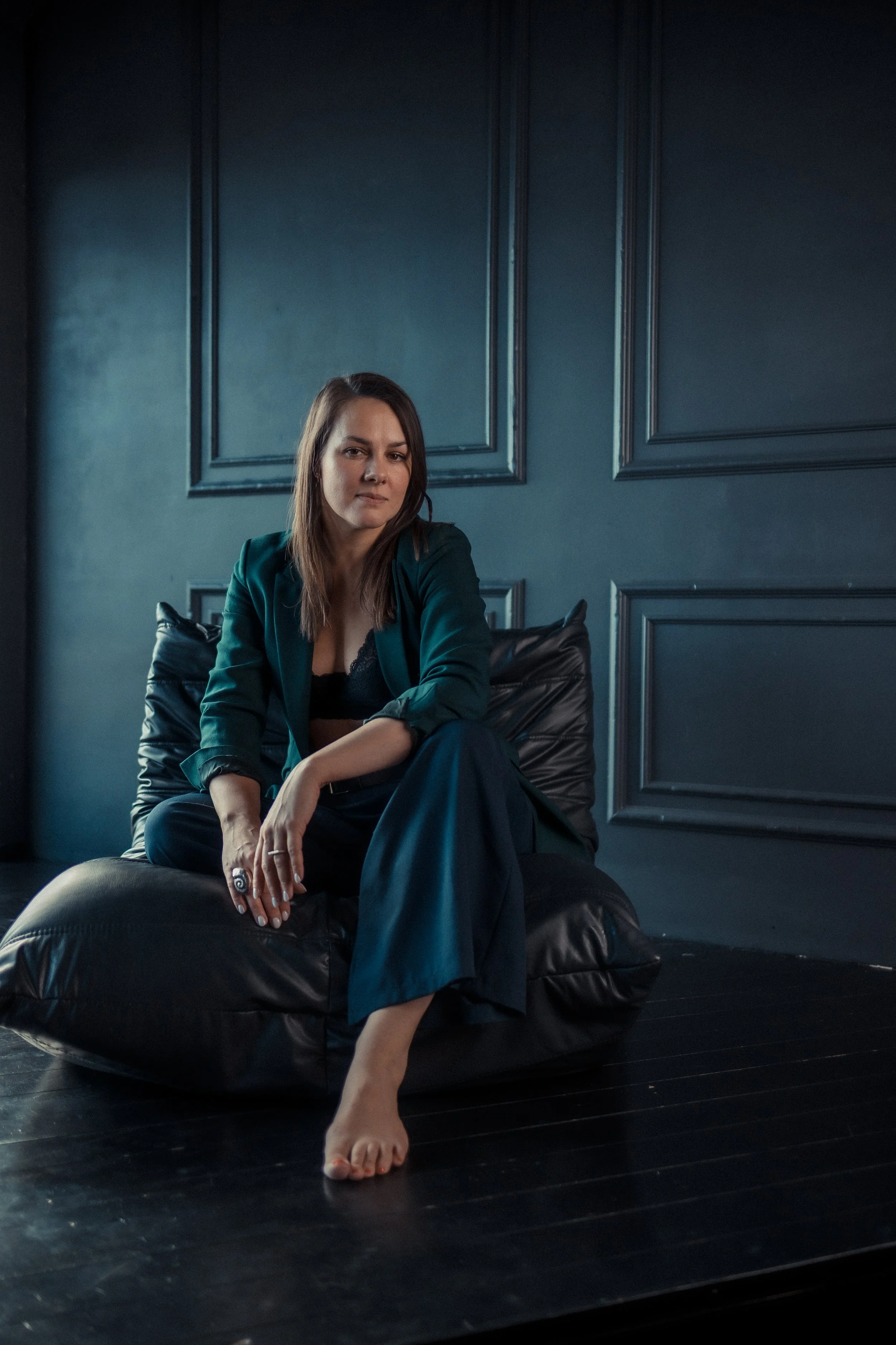 A woman sitting on a black leather couch in a dimly lit room with dark blue paneled walls, wearing a dark green blazer and black pants, with one leg crossed over the other and a ring on her finger.