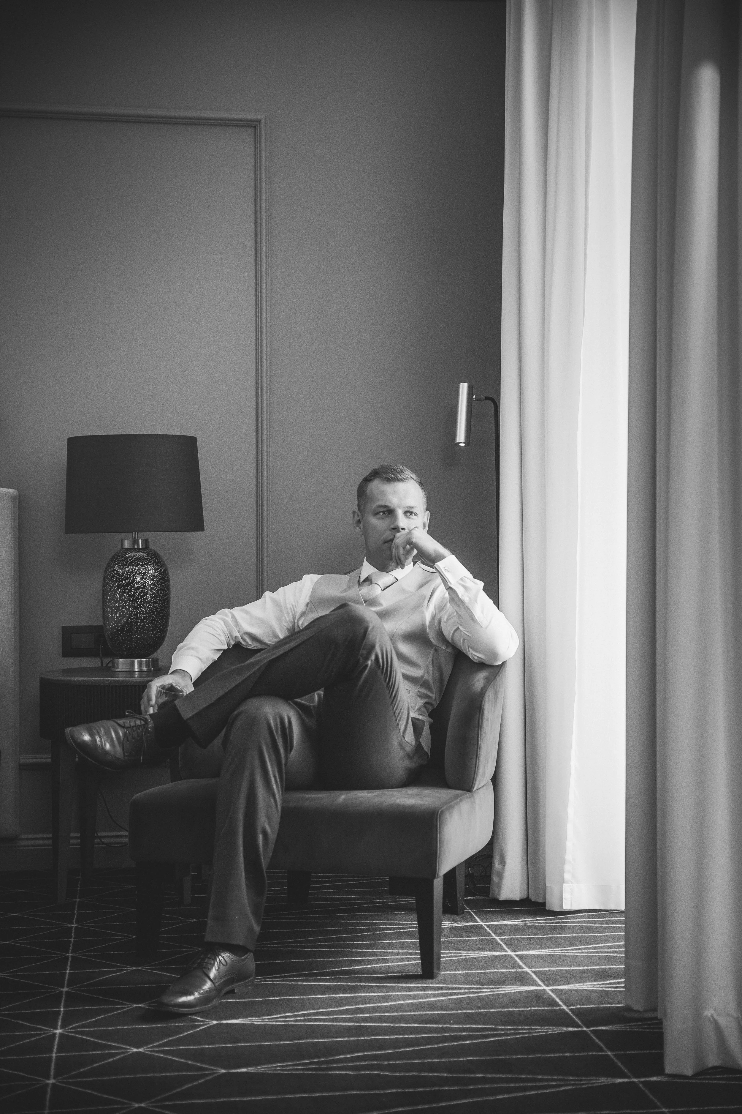 A black-and-white photo of a man sitting in an armchair in a hotel room. He is dressed in a white shirt, tie, and dark trousers, with one leg crossed over the other. The room has a side table, a lamp, curtains, and a wall-mounted reading light.