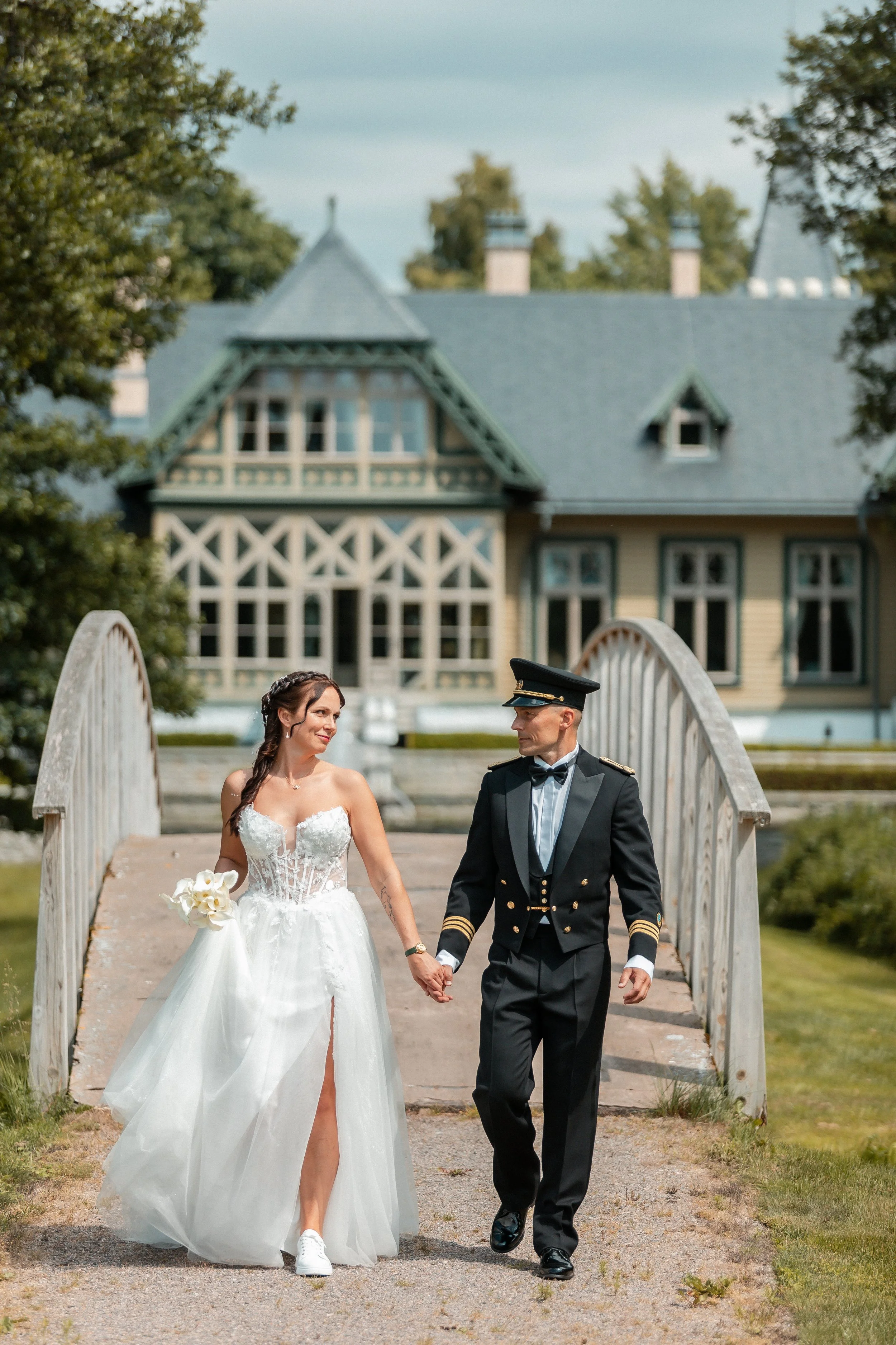 A bride and groom holding hands and walking on a bridge outdoors, with a house in the background.