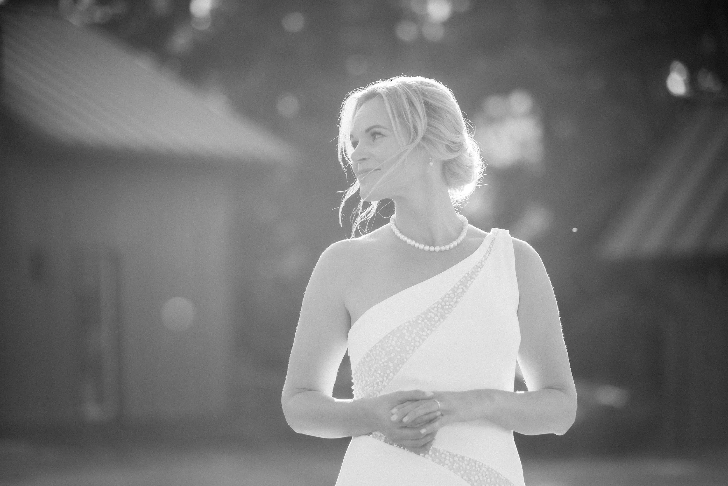A woman with light hair styled in an elegant updo wearing a white dress with embellishments and a pearl necklace, standing outdoors with soft sunlight and blurred background.