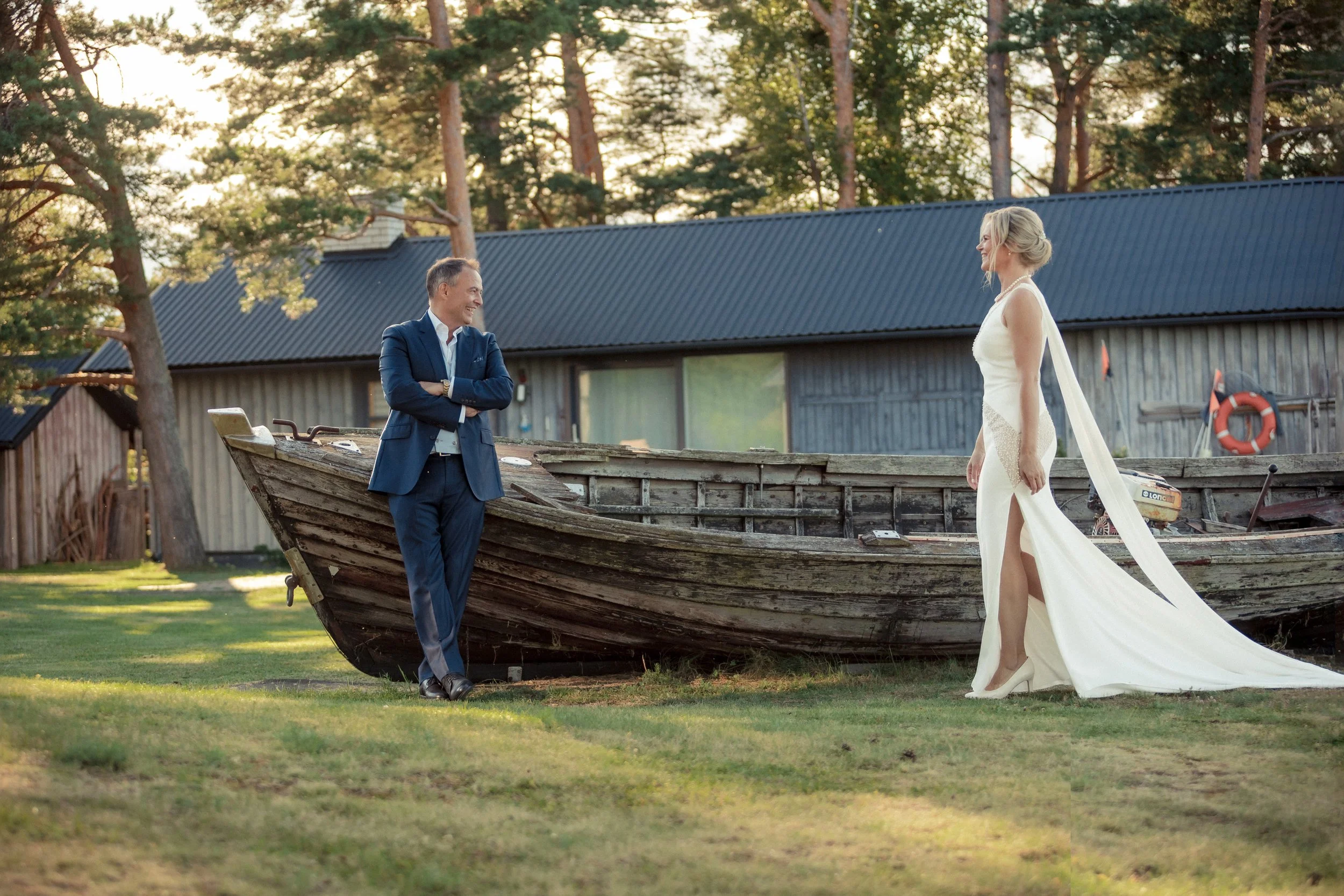 A man in a suit and a woman in a wedding dress standing by a wooden boat outdoors, with trees and a building in the background, during sunset.