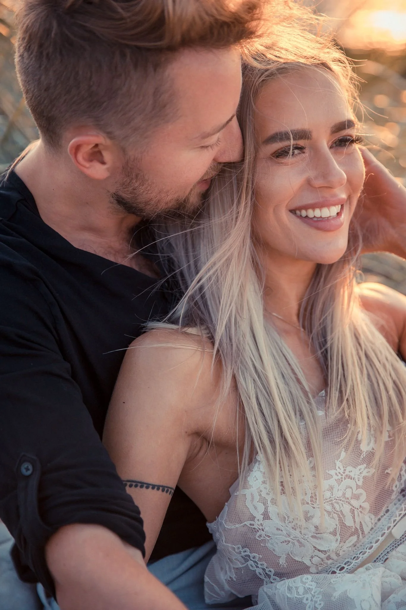 A smiling woman with long blonde hair and fair skin, wearing a white lace dress, being embraced by a man with short brown hair, a beard, and wearing a black shirt, outdoors during sunset.