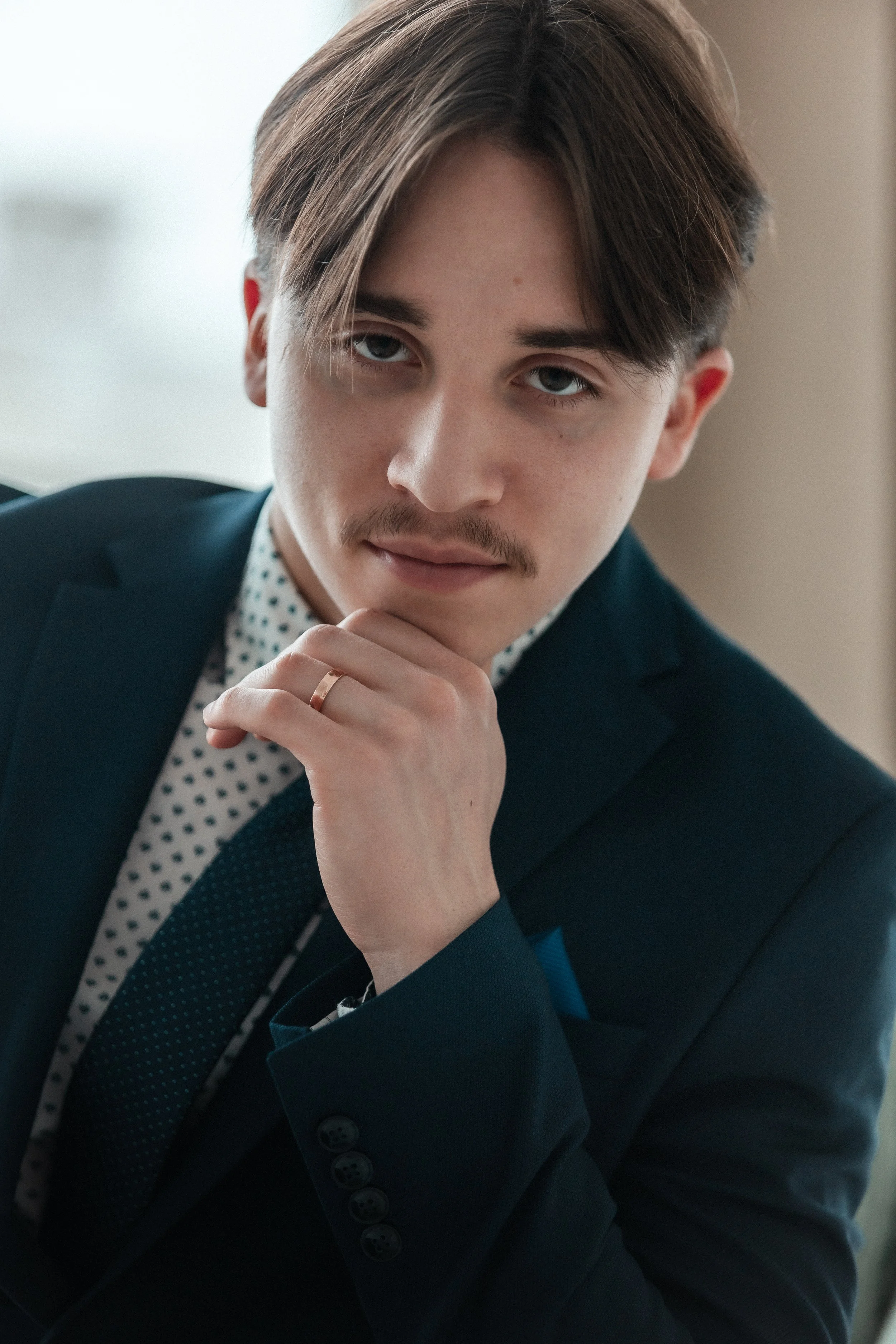 A young man with light skin, brown hair, and blue eyes, wearing a dark suit and a polka-dot shirt, is resting his chin on his hand and looking at the camera.