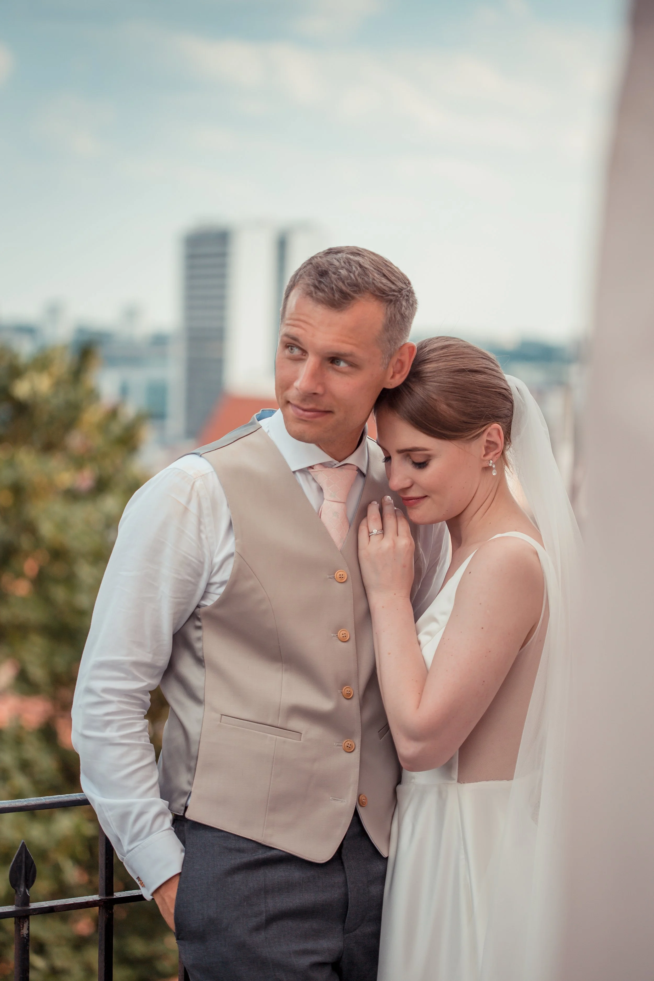 A bride and groom standing close together, with the groom looking to the side and the bride resting her head on his shoulder, outdoors with blurred city buildings in the background.