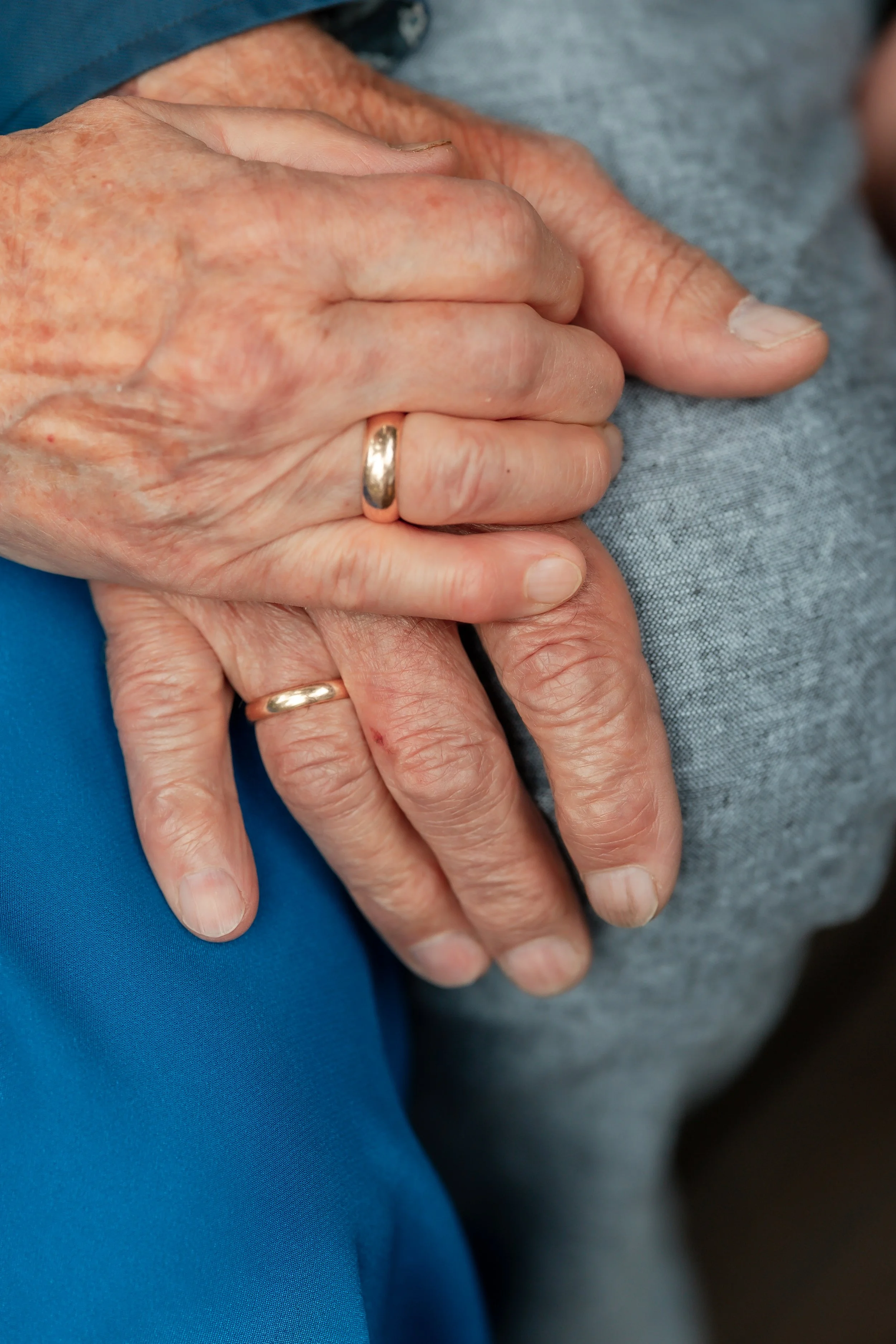 Close-up of two elderly people's hands clasped together, wearing wedding rings. One hand has a grayish nail, the other hand shows faint wrinkles. The hands rest on a gray fabric surface, with a touch of blue clothing visible in the background.