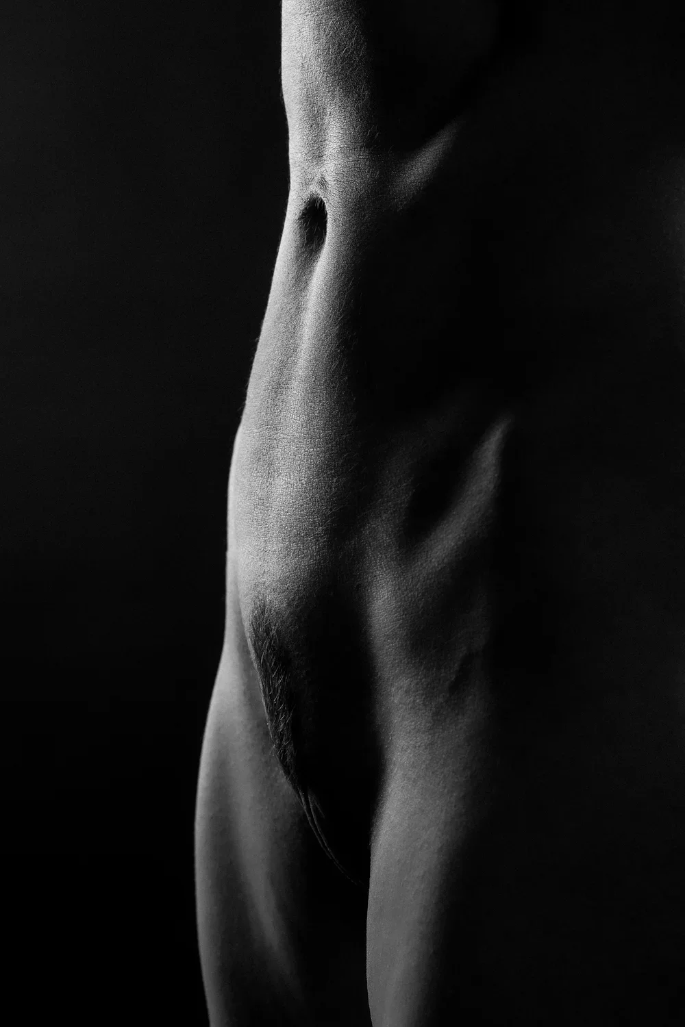 Close-up black and white photograph of a human torso, showing the abdomen, waist, and part of the thigh, with soft lighting emphasizing the skin's texture.