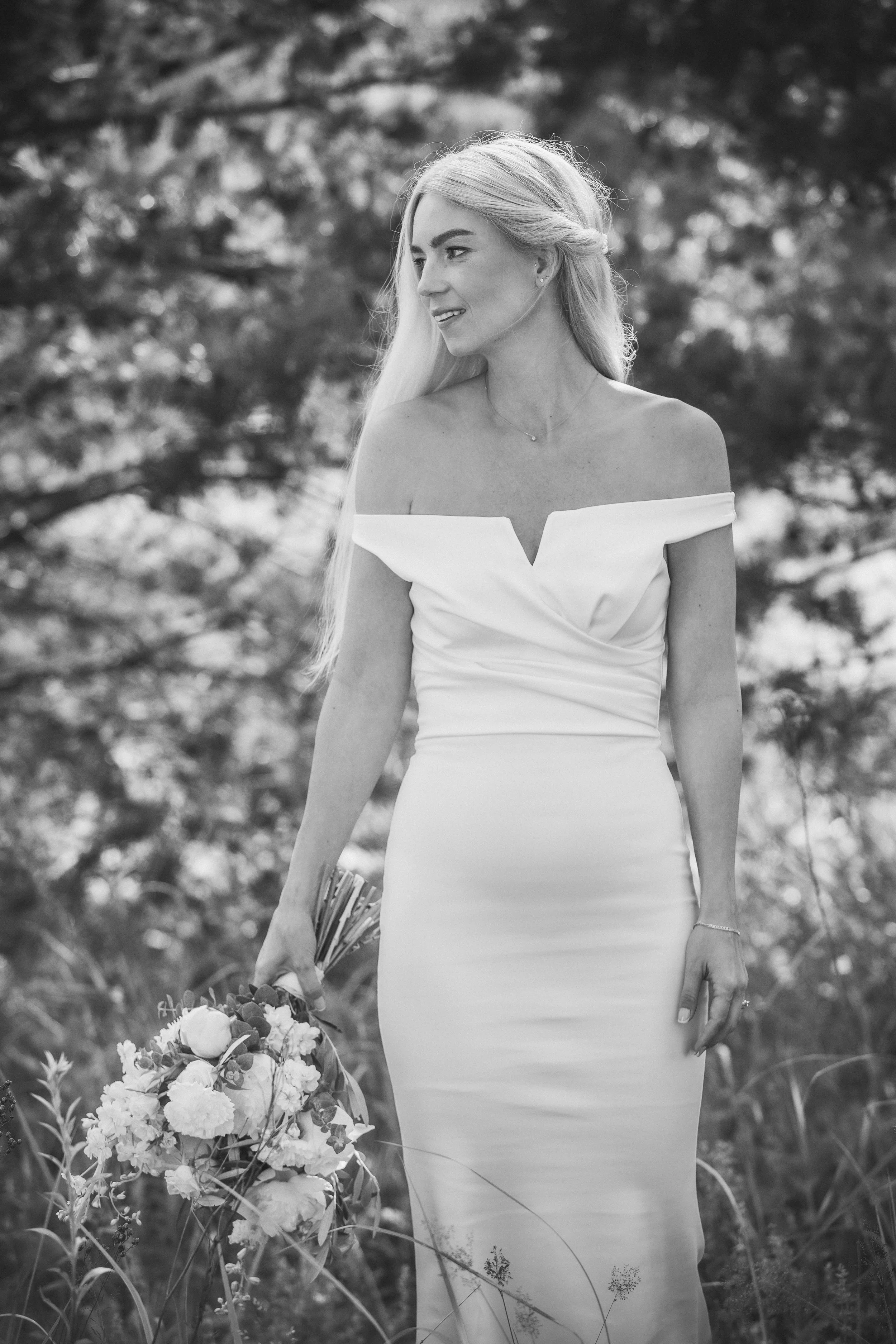 Black and white photograph of a woman in a formal off-the-shoulder dress outdoors, holding a bouquet of flowers, surrounded by natural foliage and trees.