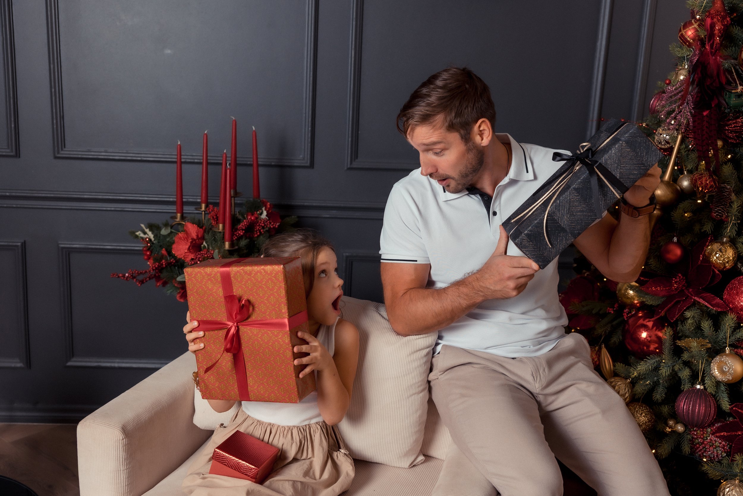 A man and a young girl with light skin and brown hair in a ponytail sitting on a beige sofa near a Christmas tree. The girl is holding a large wrapped gift with red ribbon, and the man is giving her a smaller gift with black wrapping and gold ribbon.