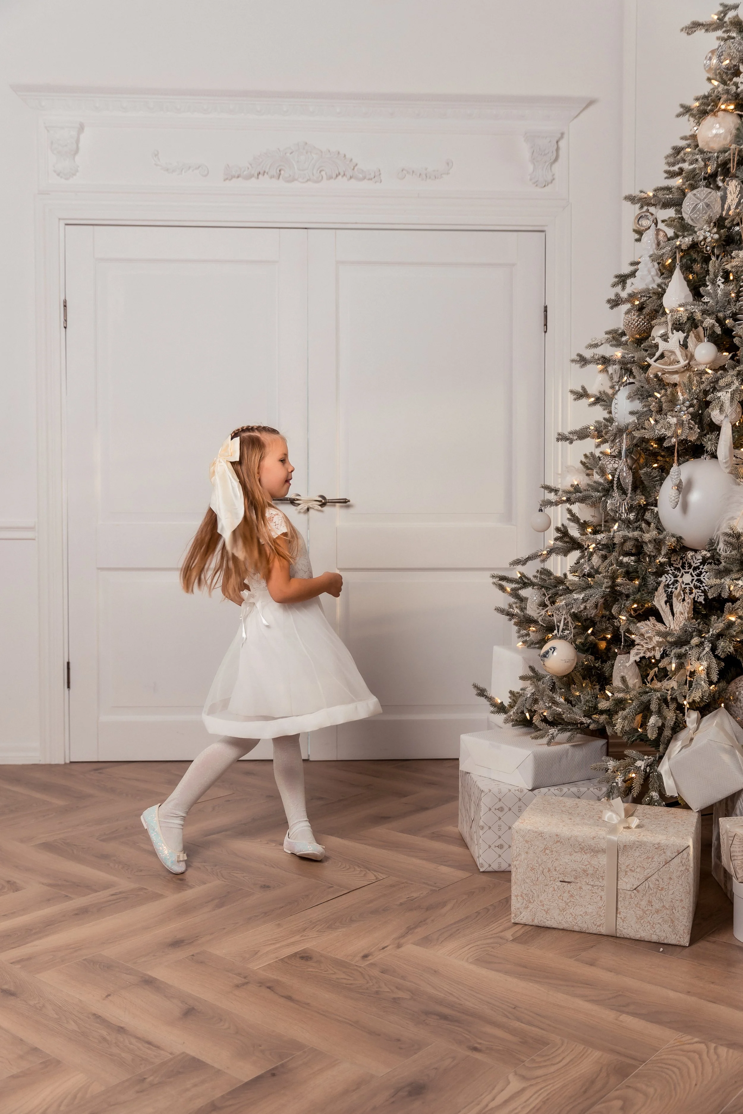 A young girl in a white dress and tights is dancing or playing near a decorated Christmas tree with wrapped presents underneath. The room has light-colored walls and wooden flooring.