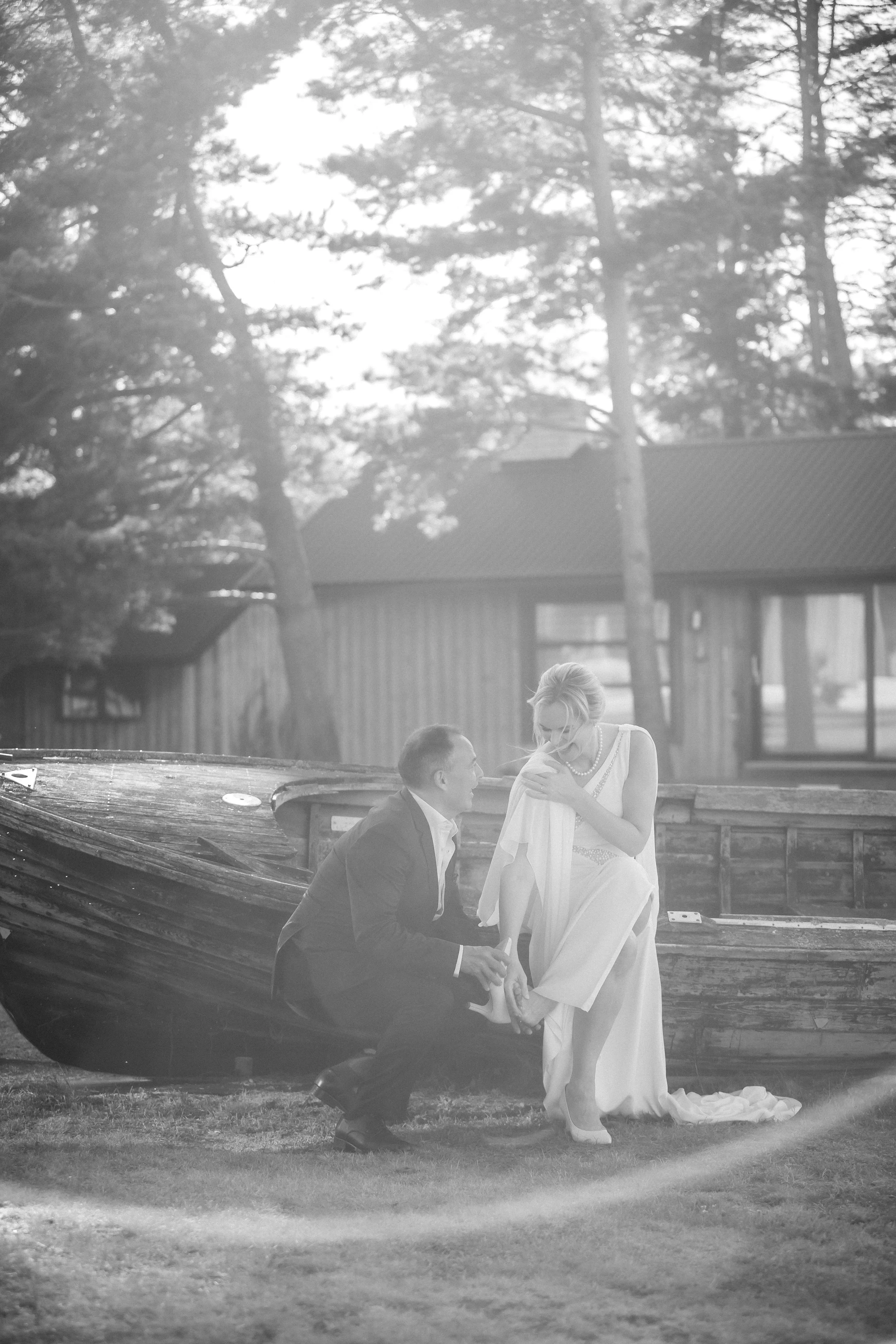 A black and white photo of a man kneeling on one knee, proposing to a woman who is holding a white dress, by a boat outdoors with trees in the background.