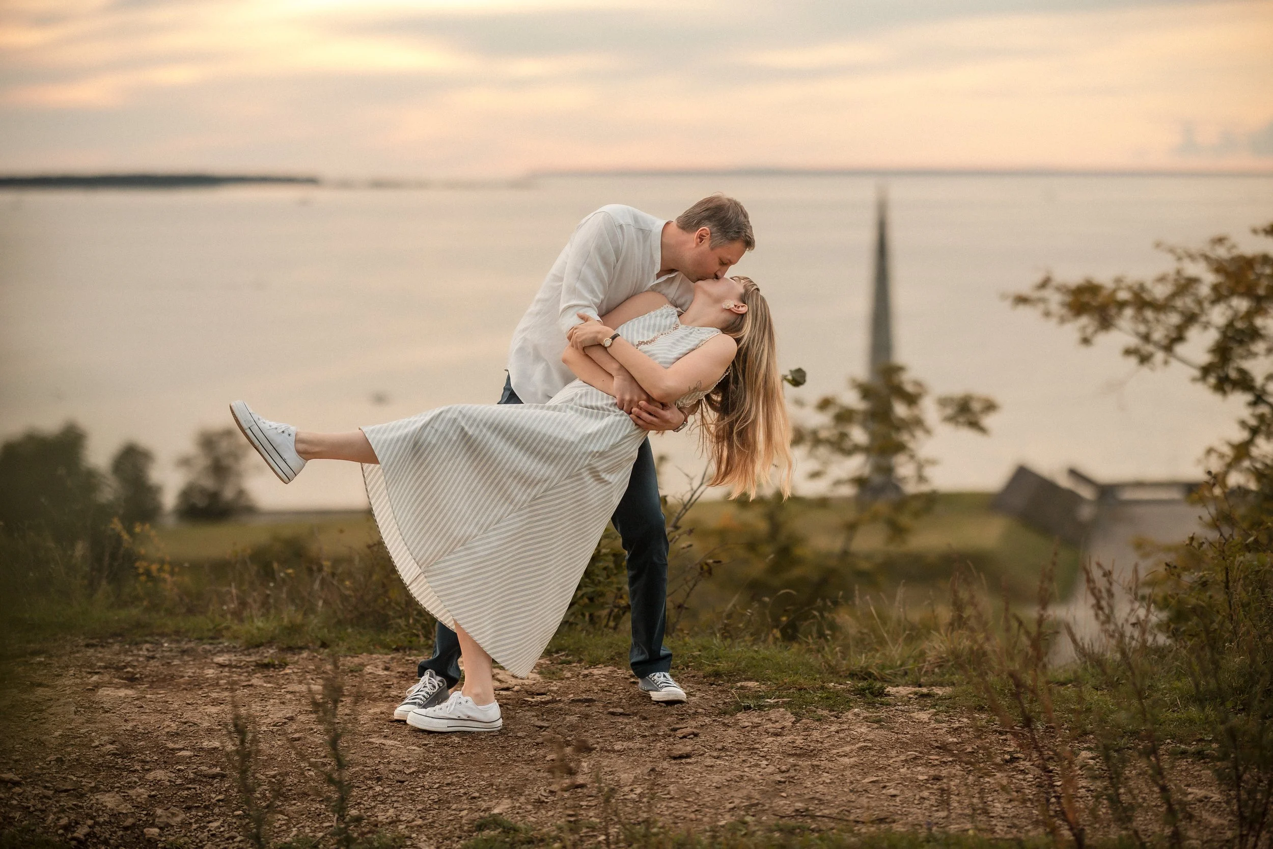A man is holding a woman in a dip position outdoors near a body of water during sunset, with trees and a cloudy sky in the background.