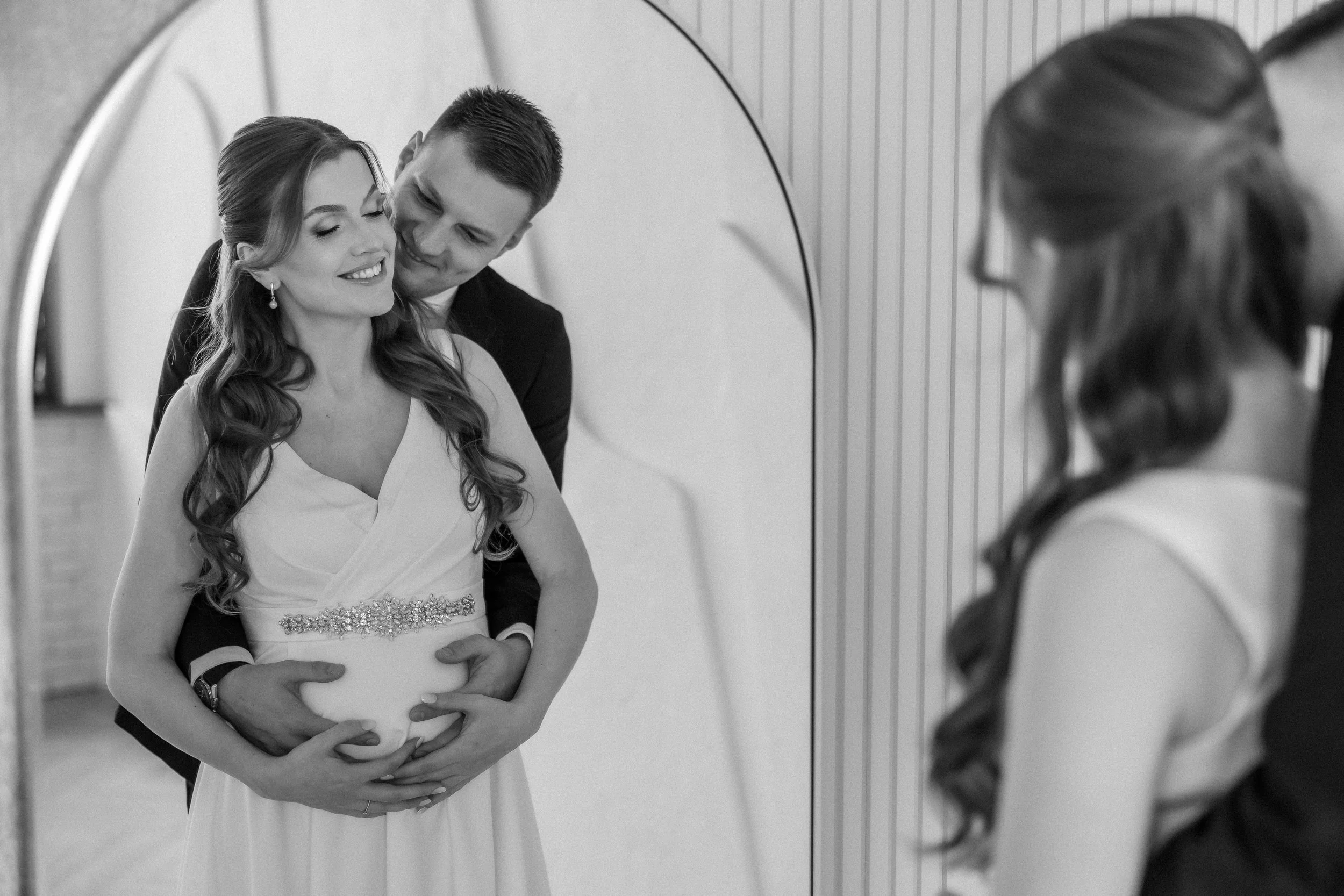 A bride and groom pose for a photograph in front of a mirror, with the groom standing behind the bride holding her belly, both smiling. The bride wears a white wedding dress with a decorative waistband, and the groom wears a dark suit. The photo is i