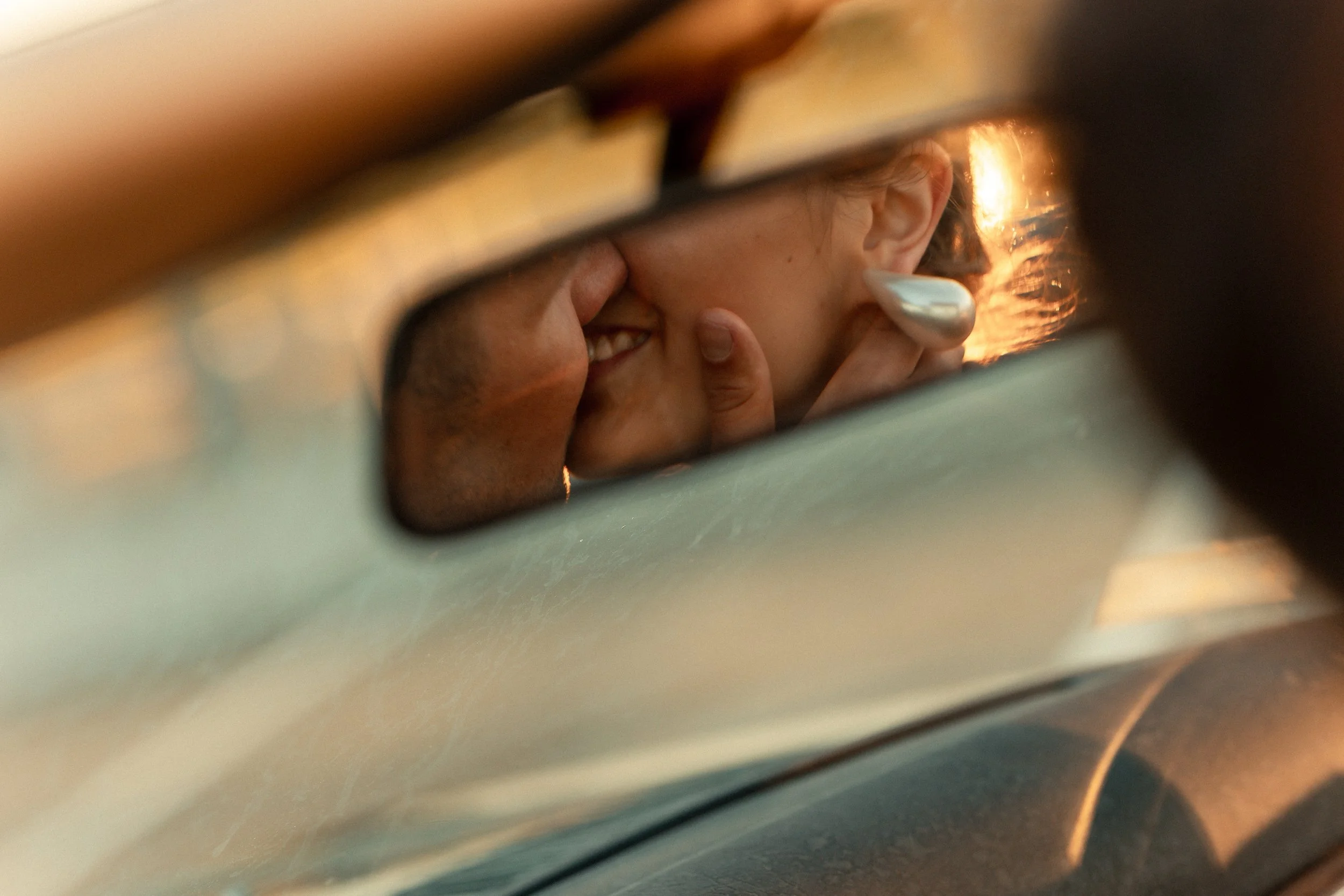 The photo, taken through a gap, shows a person with light brown hair until the neck, smiling and covering their mouth with their hand, with part of their face visible and illuminated by warm sunlight behind.