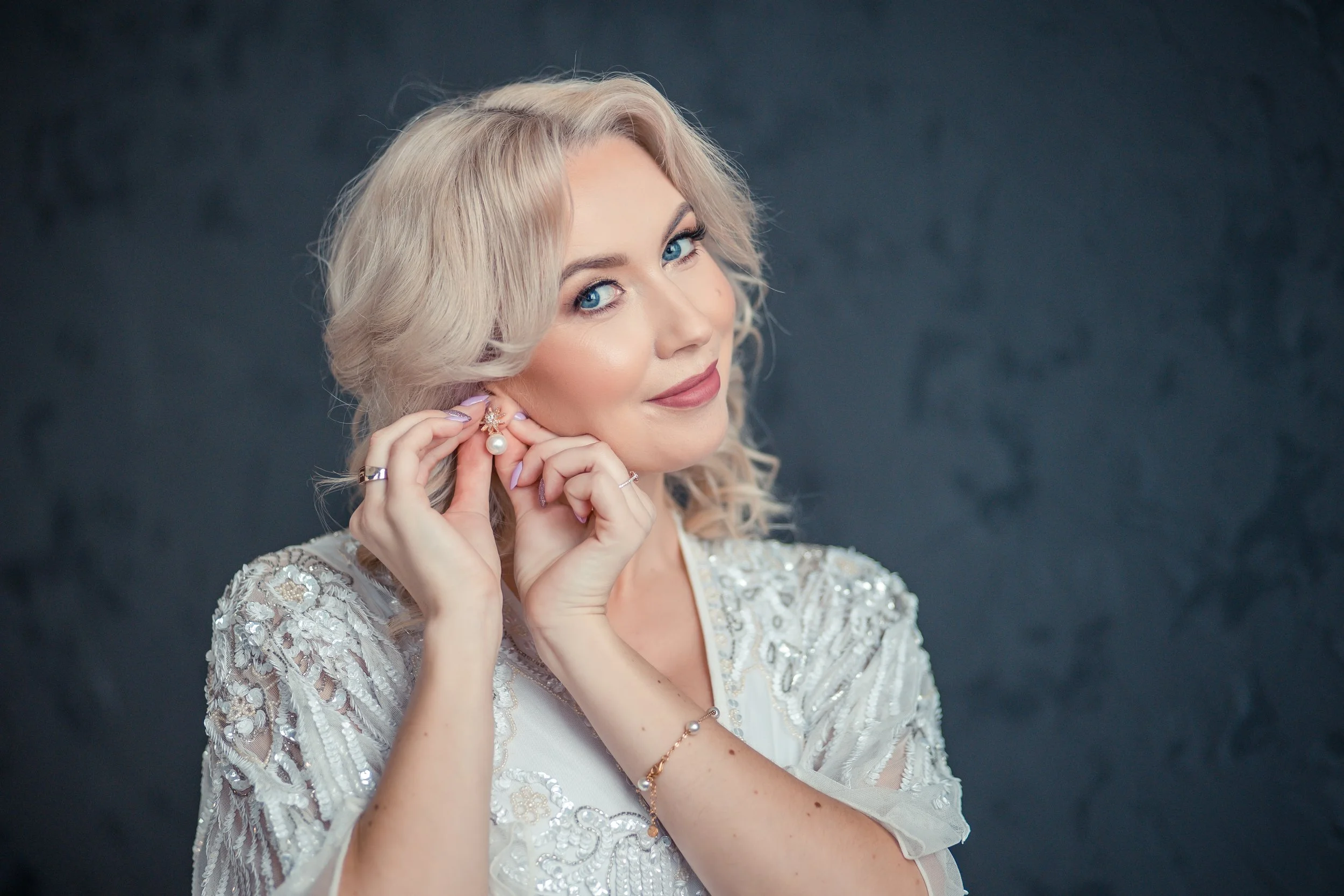 A woman with blond, wavy hair, wearing a white lace dress with intricate silver embroidery, is holding a pearl earring close to her ear and smiling softly against a dark background.