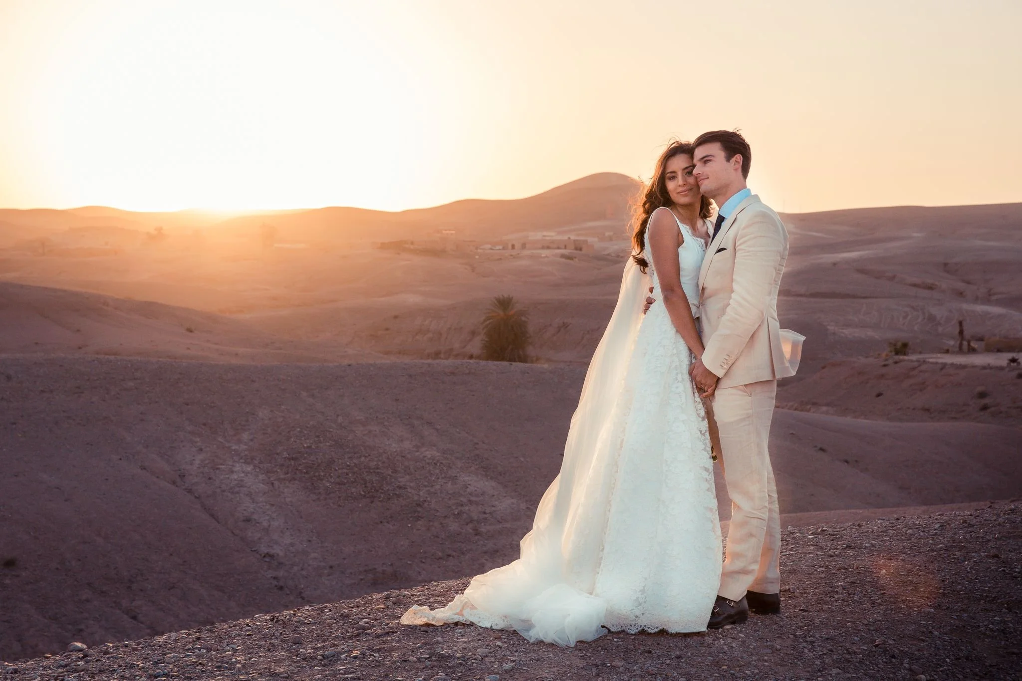 A couple dressed in wedding attire standing close together in a desert landscape during sunset.