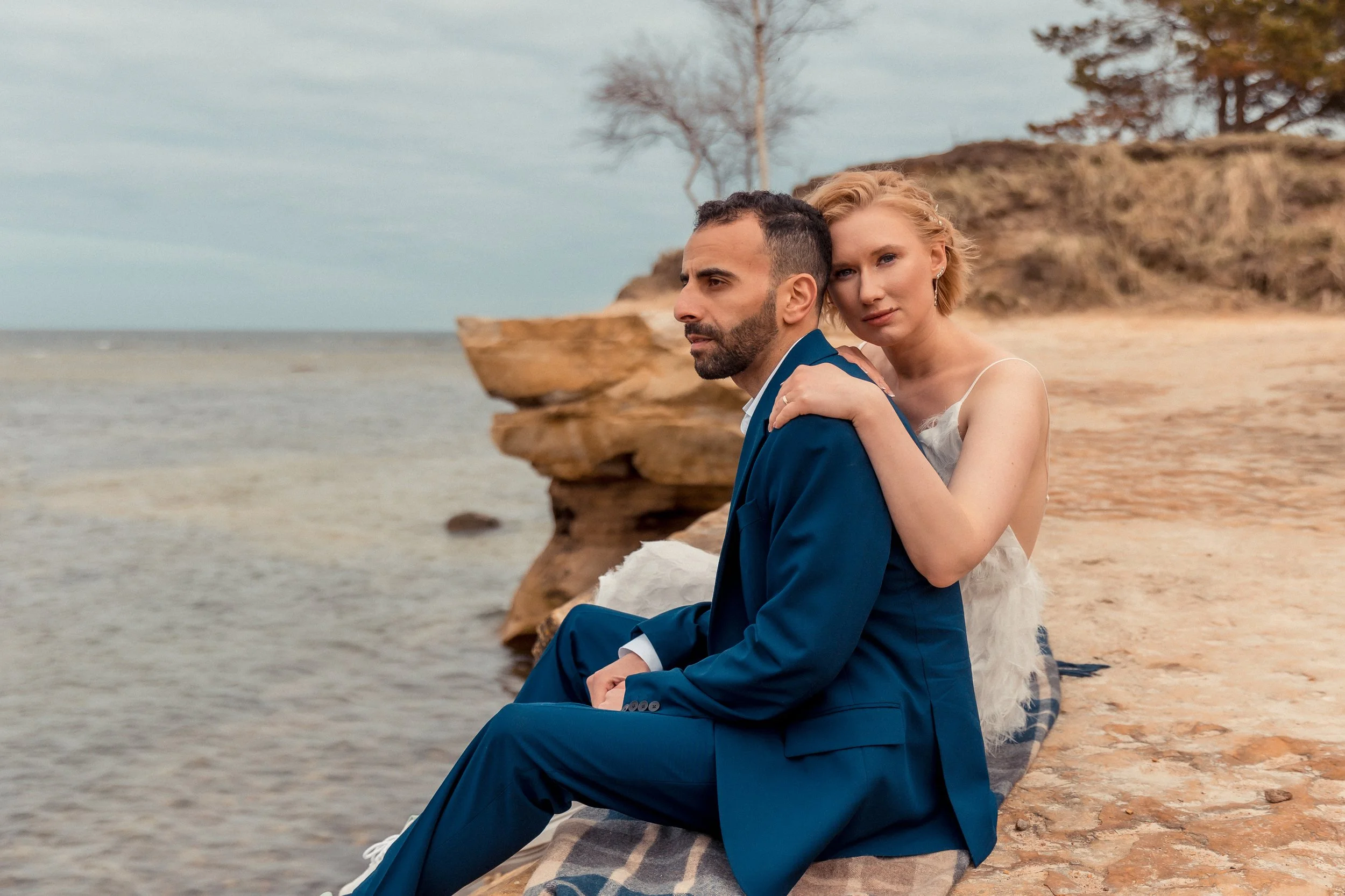 A woman in a wedding dress hugging a man in a blue suit sitting on a beach by the water with driftwood and trees in the background.