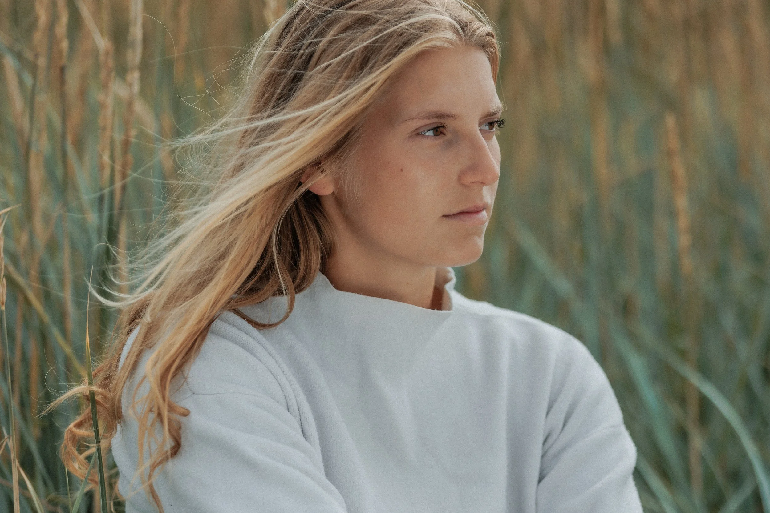 A young woman with long, wavy blonde hair wearing a white shirt, standing outdoors near tall grasses, looking to the right with a thoughtful expression.