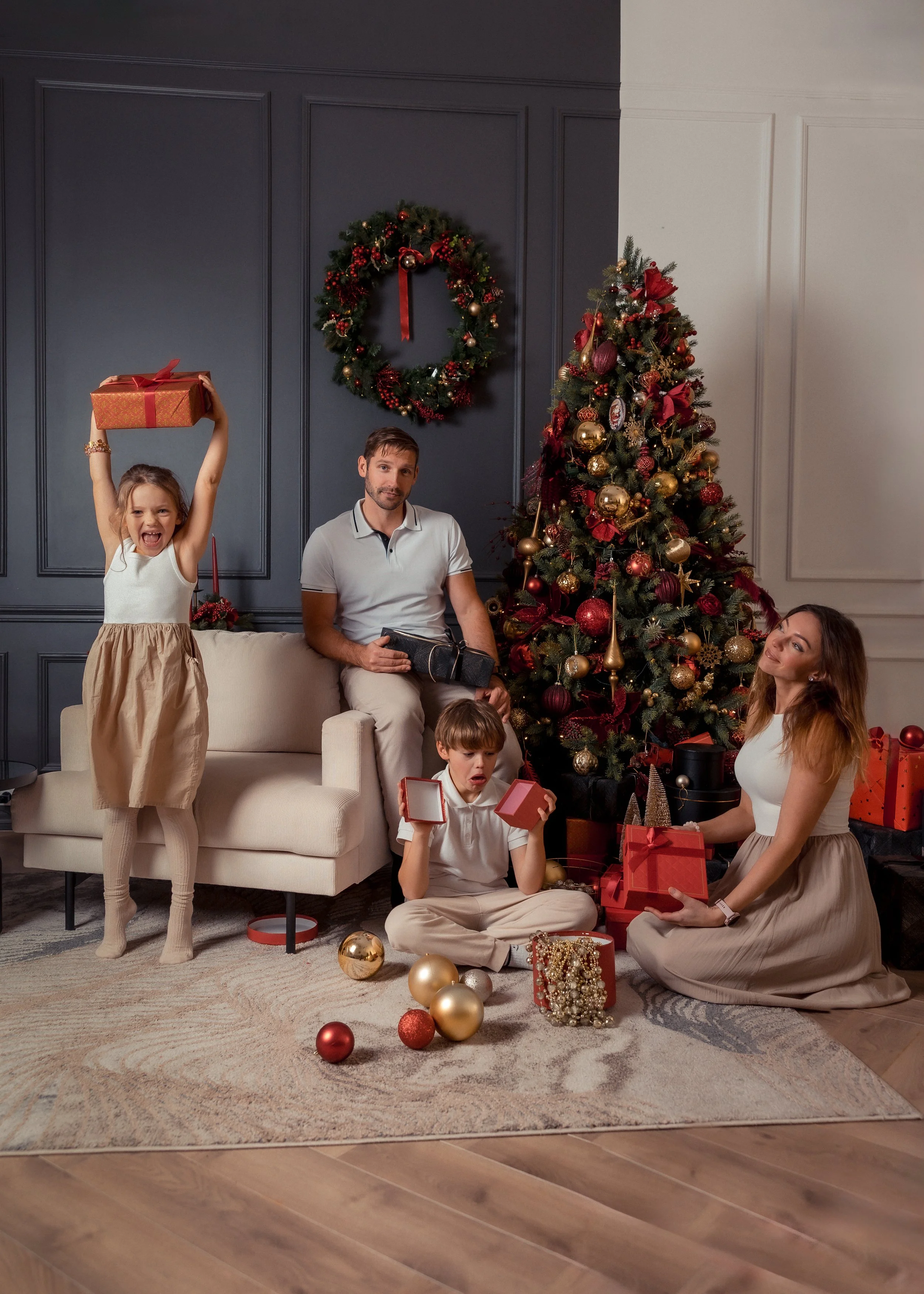 A family celebrating Christmas in a living room decorated with a Christmas tree, ornaments, and wreath on the wall. A girl is joyfully raising a wrapped gift, a man is sitting with a present on his lap, a woman is kneeling with a gift, and two boys a