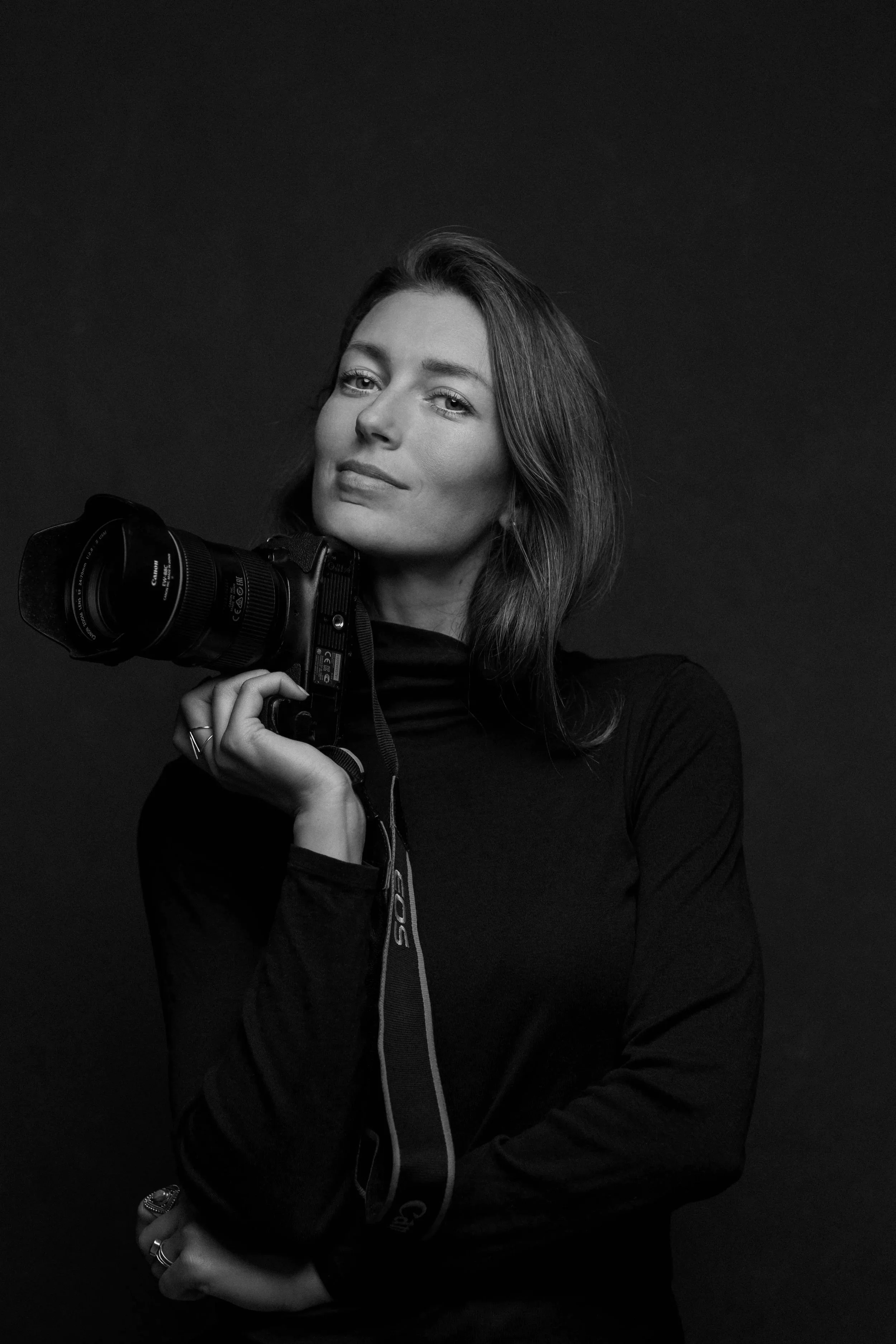A black and white portrait of a woman holding a camera with her right hand, wearing a long-sleeve top and several rings, against a dark background.