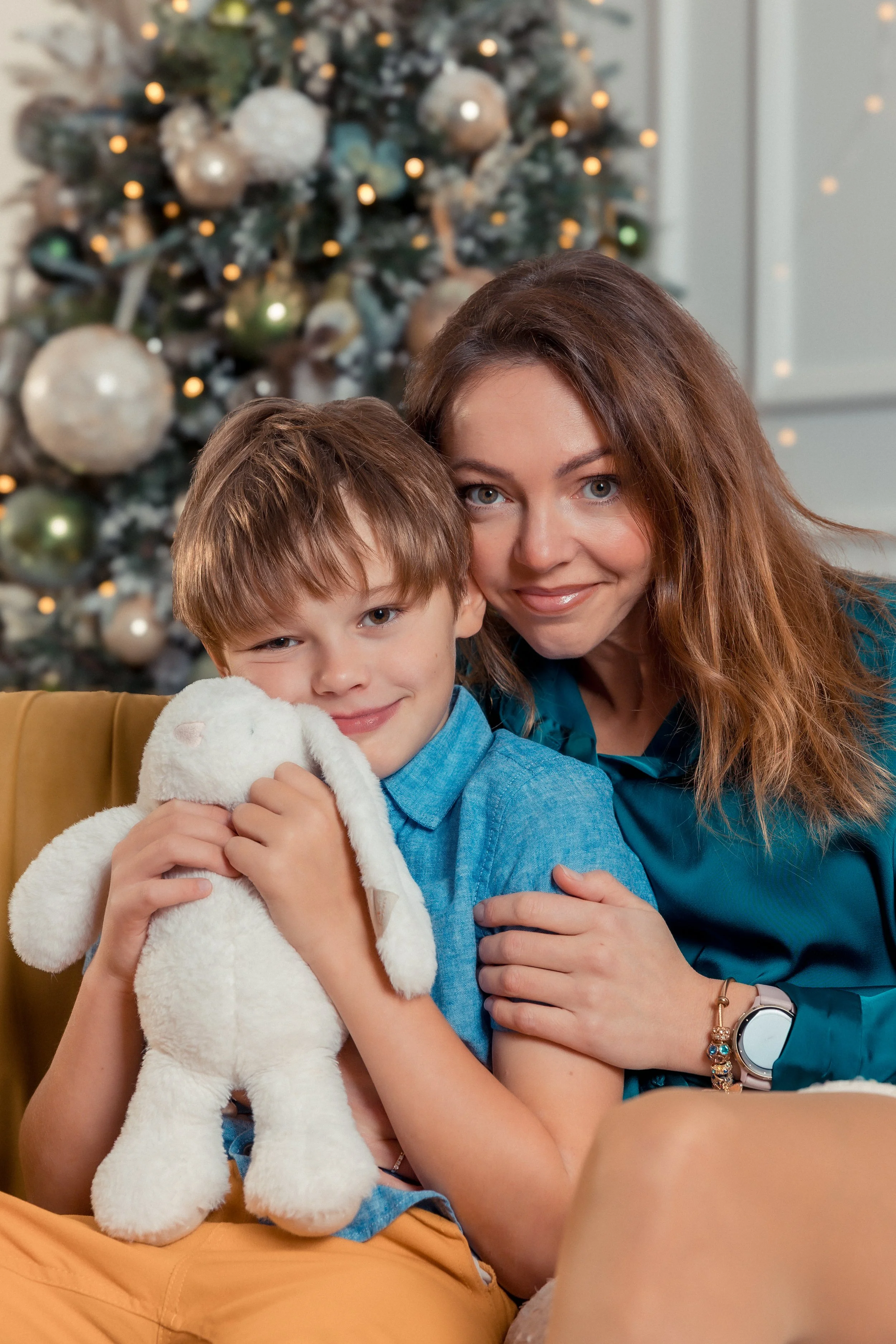 A woman with long brown hair and a young boy with brown hair and blue eyes sitting in front of a decorated Christmas tree, holding a white stuffed bunny, both smiling at the camera.