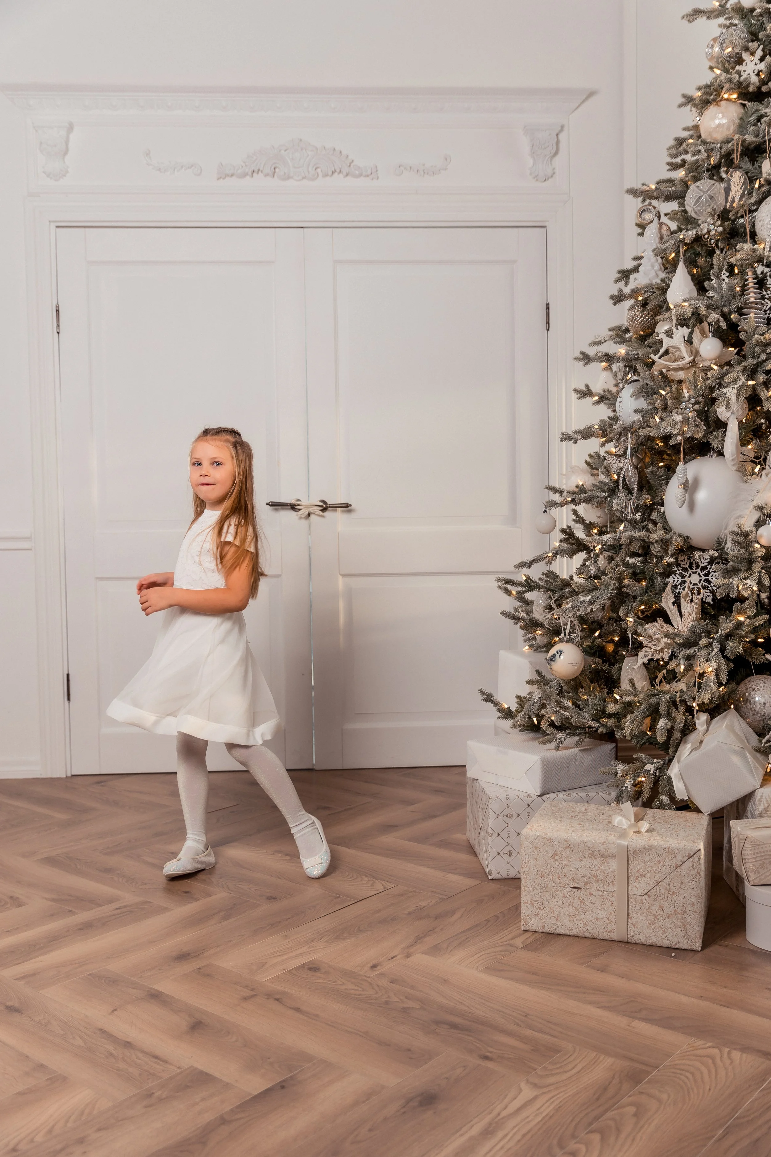 A young girl in a white dress and tights is dancing in front of a white door next to a decorated Christmas tree with presents underneath.